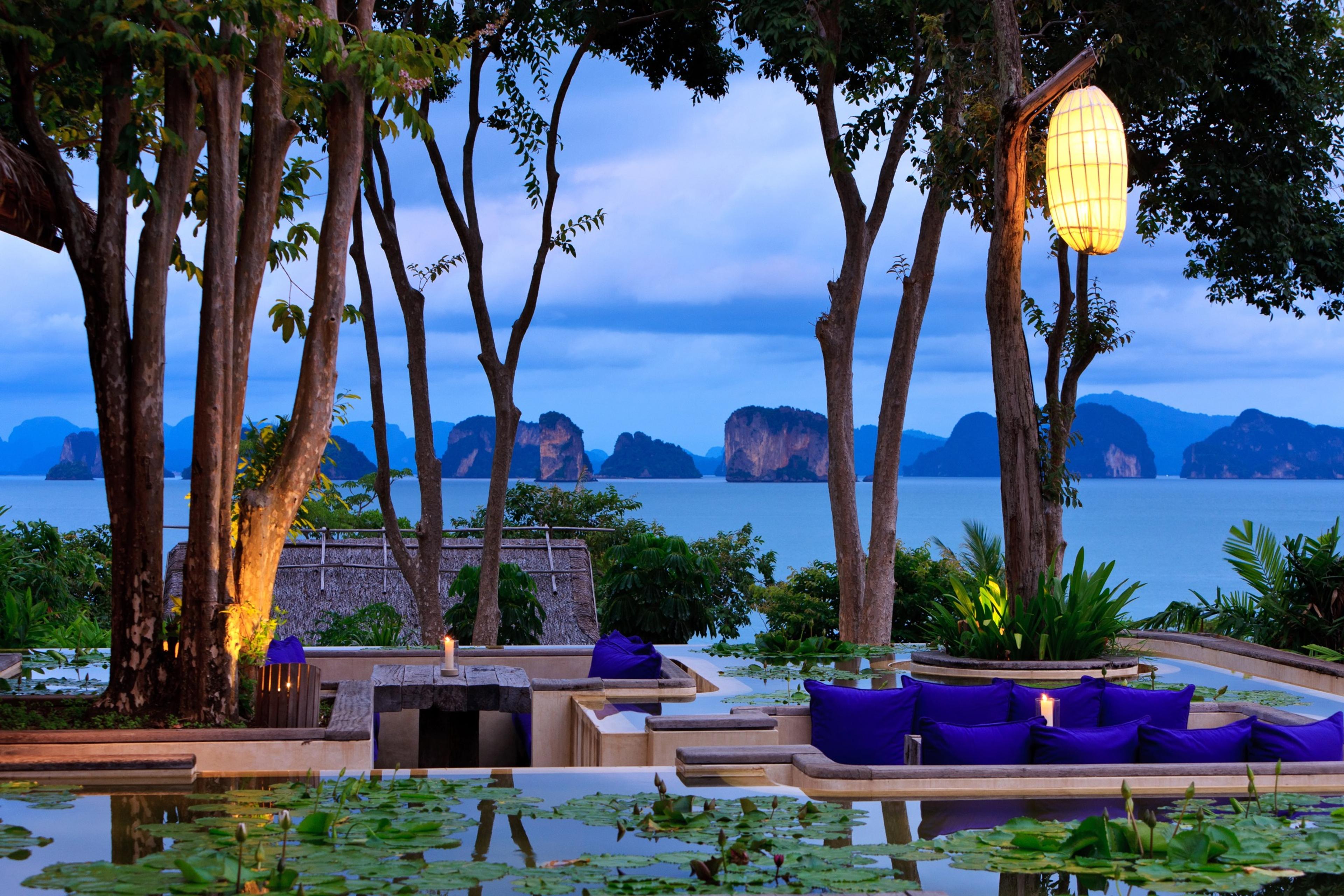 An outdoor lounge with blue cushioned seating beside a lily pond, lit by a paper lantern among trees, overlooking a bay dotted with rocky islands at dusk.