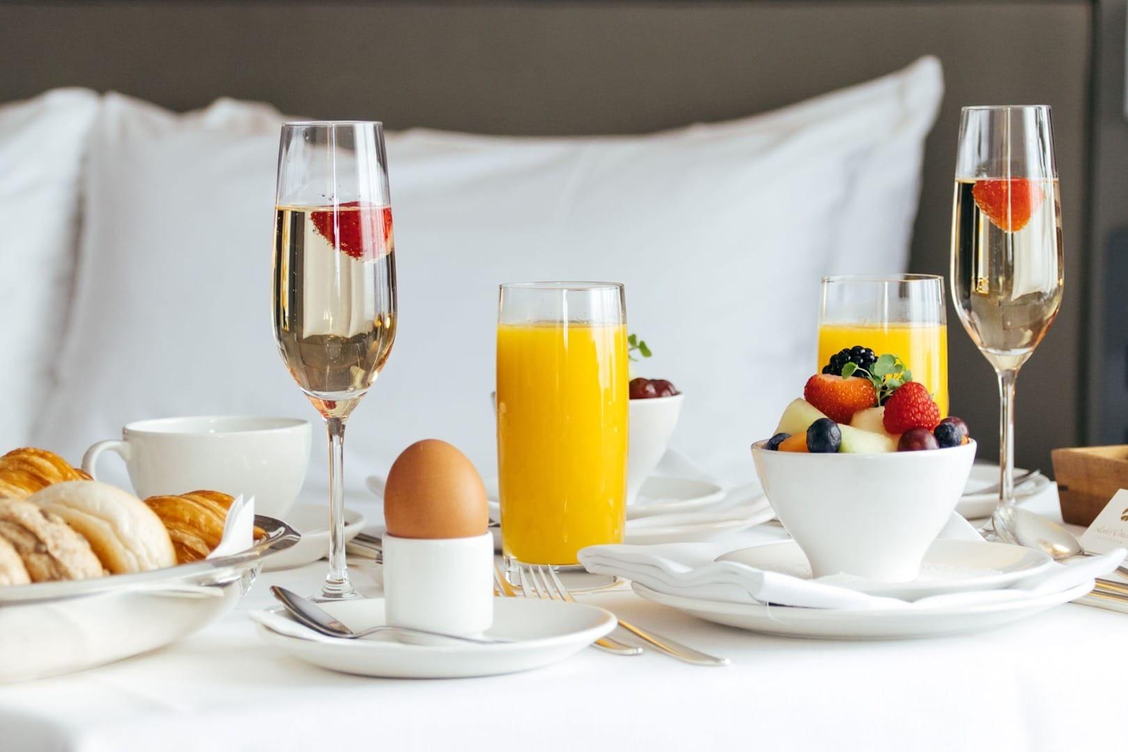 A breakfast tray on a bed with croissants, a soft-boiled egg, orange juice, a bowl of fruit, and two glasses of sparkling wine with strawberries.