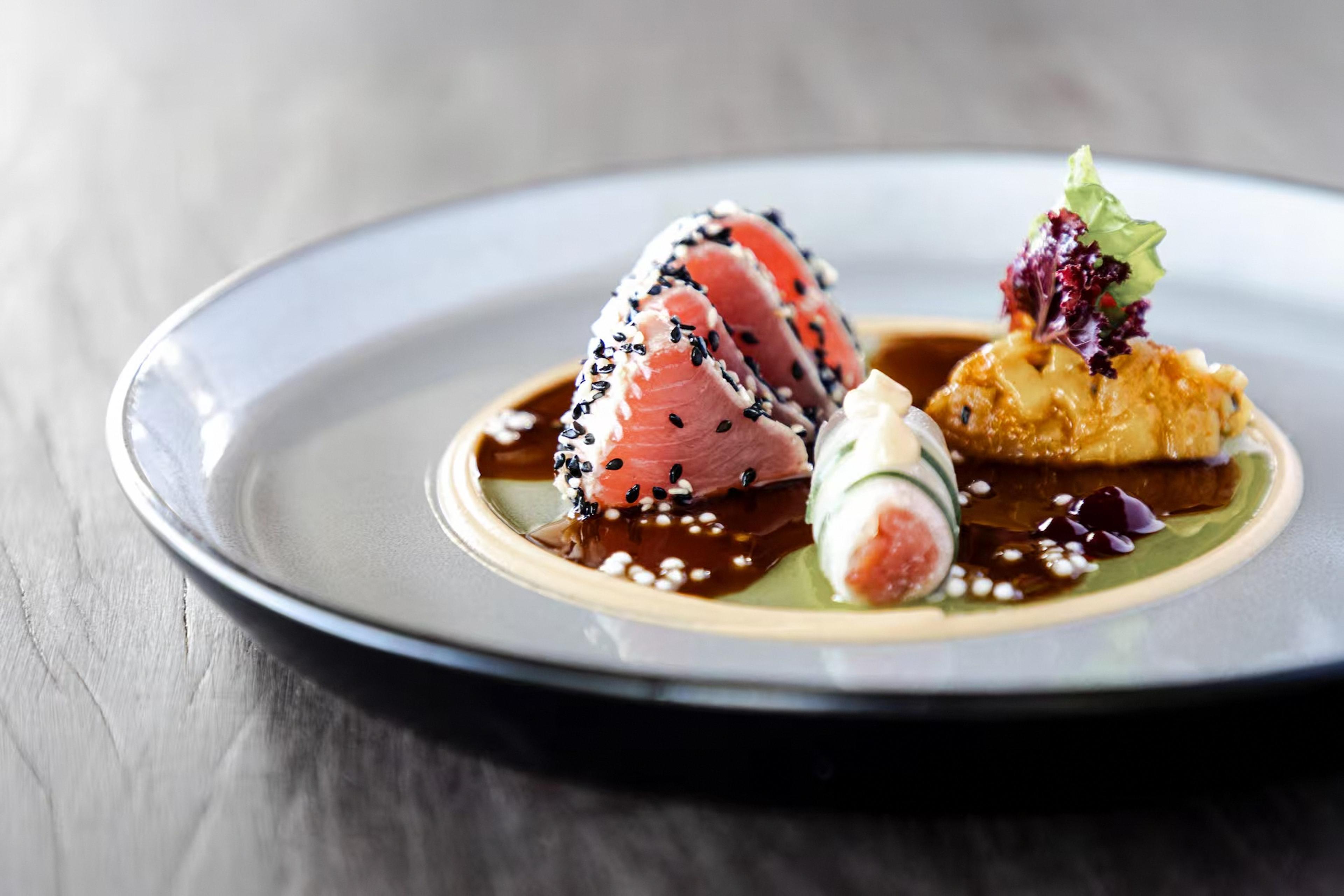 Sesame crusted seared tuna slices with cucumber and sea urchin in dark sauce on a white plate.