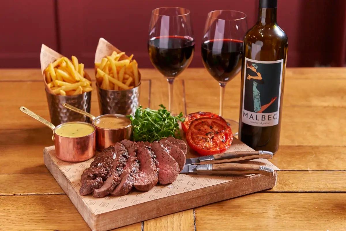 Sliced steak on a wooden board with two small copper pots of sauce, chips in paper cones, watercress and a grilled tomato half, beside two glasses of red wine and a bottle of Malbec.