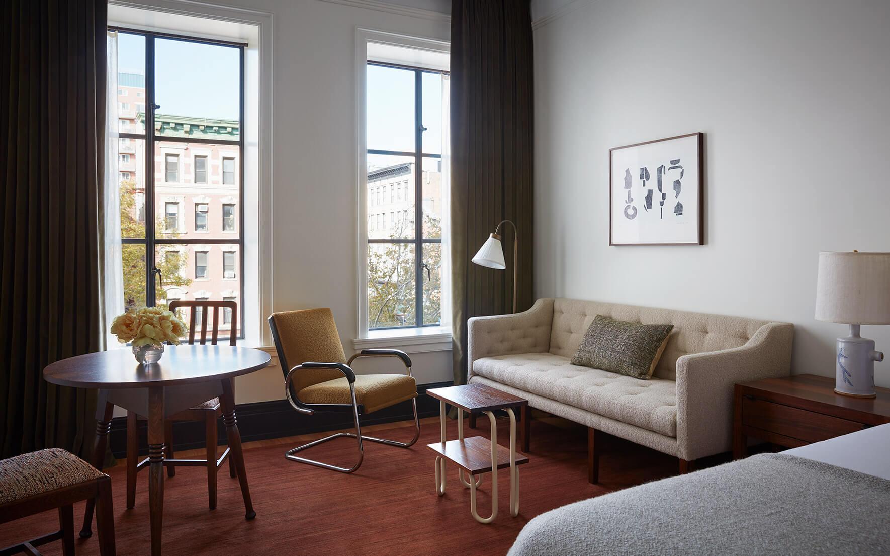 A hotel sitting room with a round table and armchair by two tall windows showing other buildings, a beige sofa against the opposite wall, and a framed print above it.