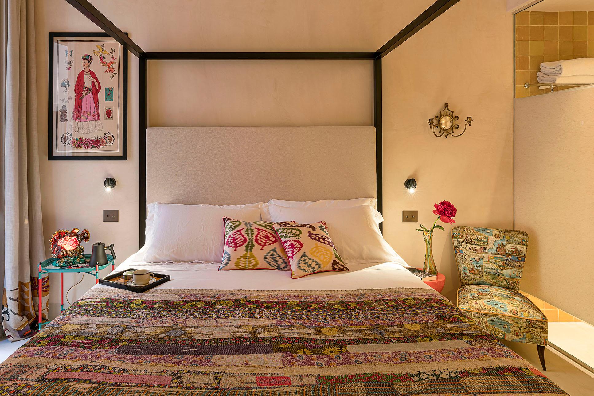 A four poster bed with a taupe headboard and patterned cushions, a botanical print on the wall, a patterned armchair, and a tiled ensuite visible beyond.