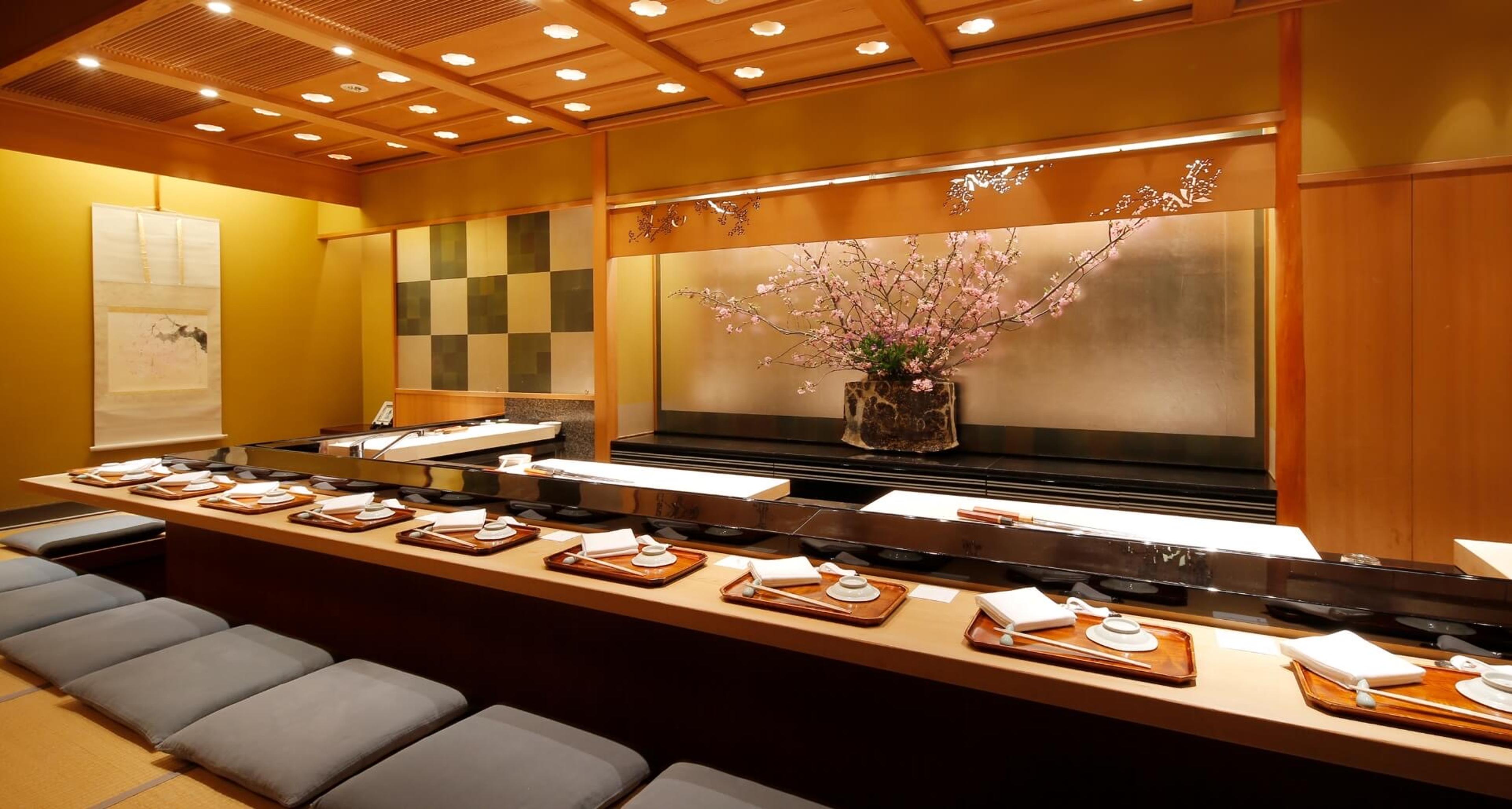 A pale wooden sushi counter laid with trays, a cherry-blossom arrangement on the wall behind