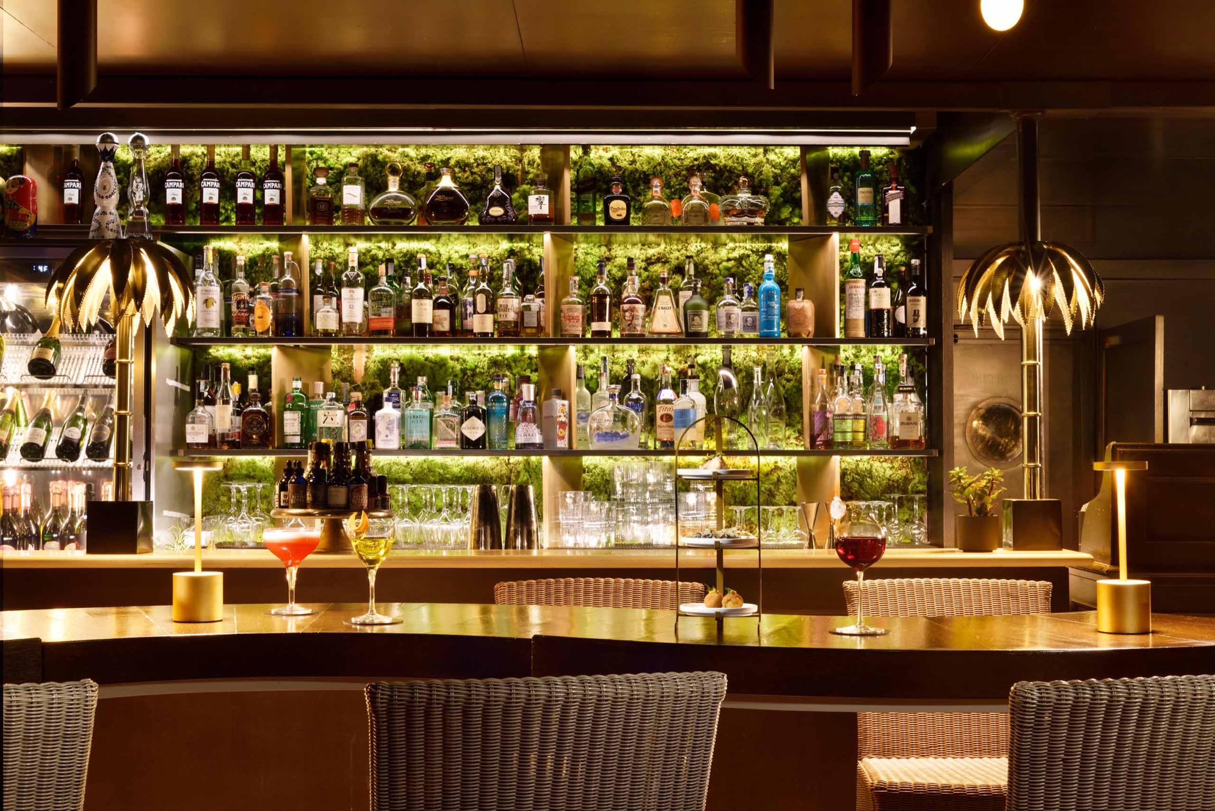 A bar with backlit shelves of bottles behind the counter, two cocktails on the bar, a cake stand of small pastries at the centre and wicker chairs in front.