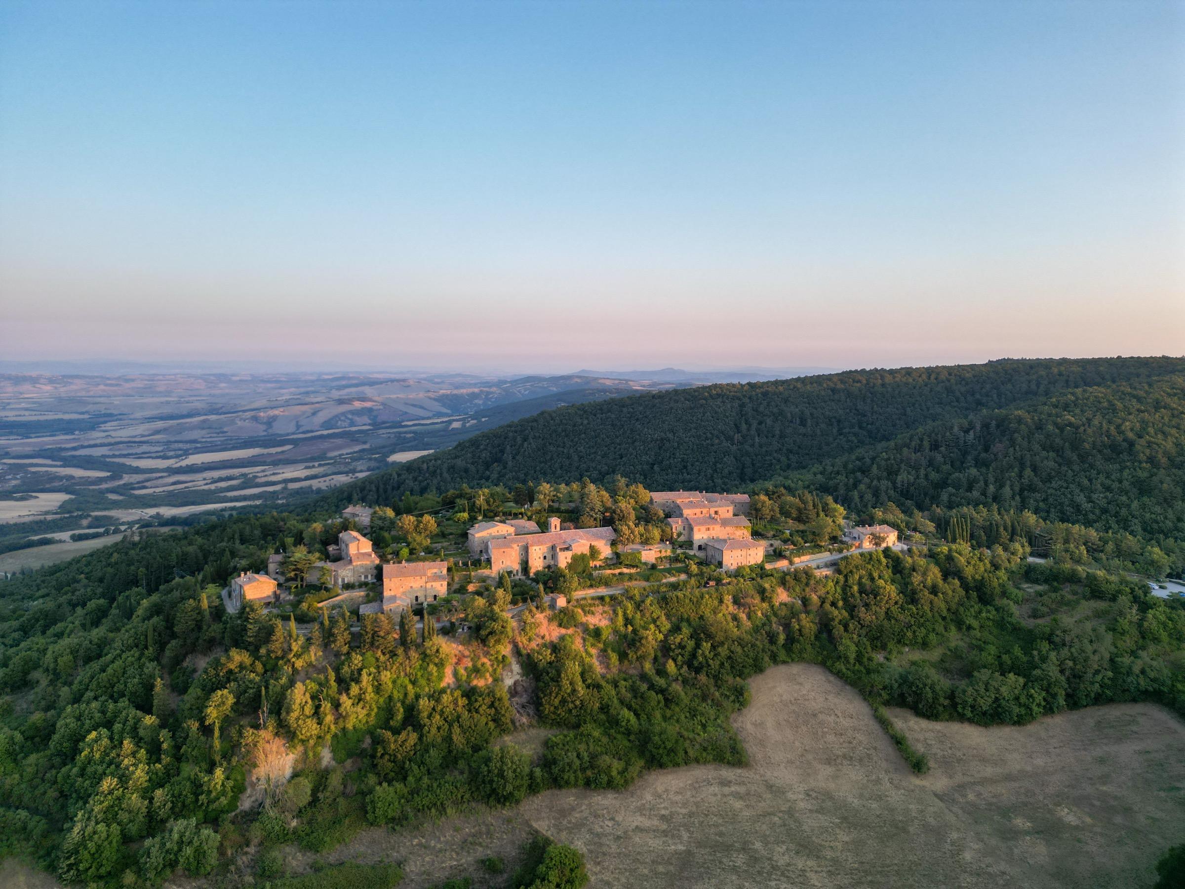 An aerial view at sunrise of a hilltop hamlet of stone buildings surrounded by forest, with a wide valley and hills stretching to the horizon.