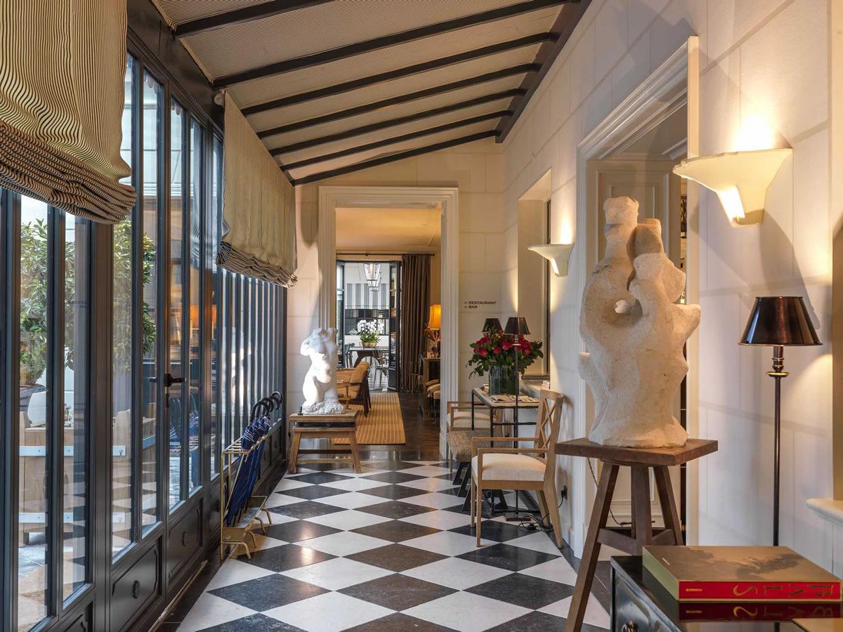A glass-walled corridor with a vaulted timber ceiling, a black and white checkerboard floor, and stone busts on wooden stands, leading past a window onto a planted courtyard.