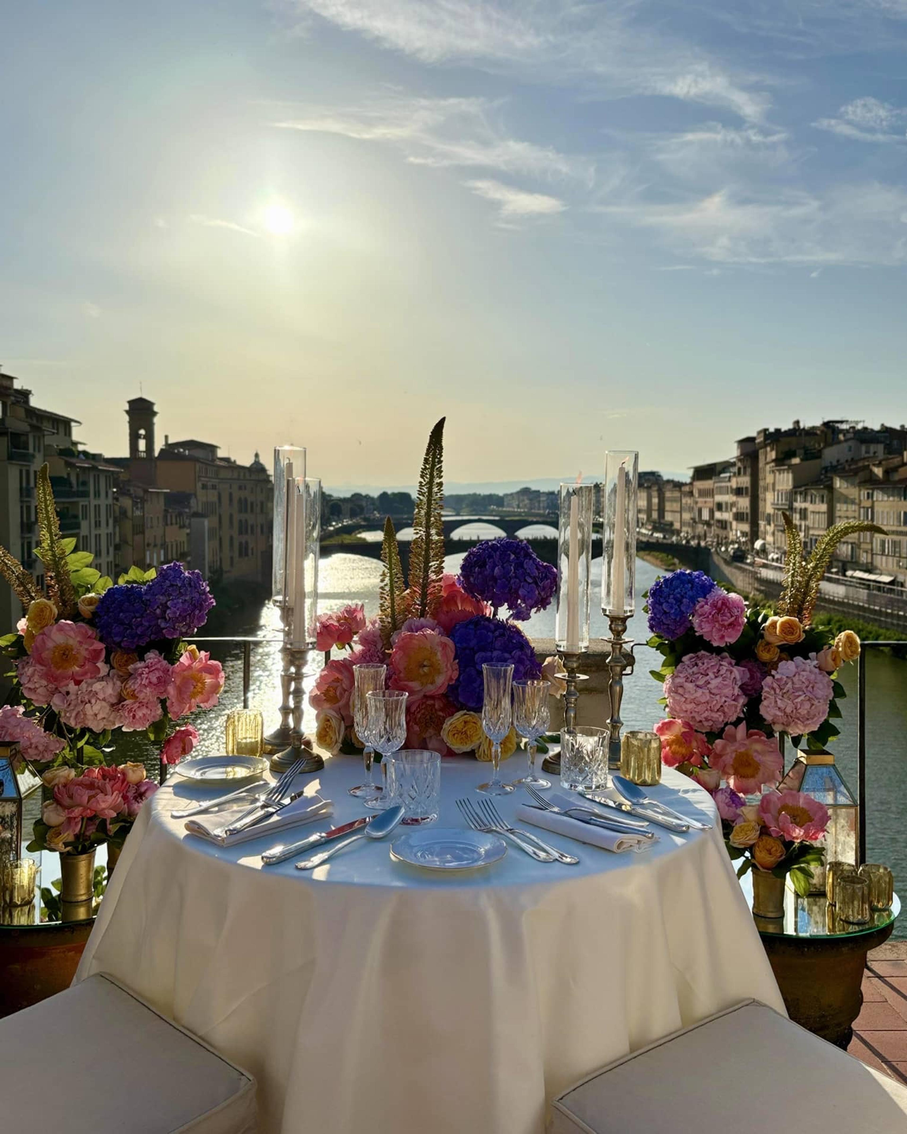 A table set for two with flowers and tall candles on a riverside terrace, looking along the Arno past its bridges at sunset.