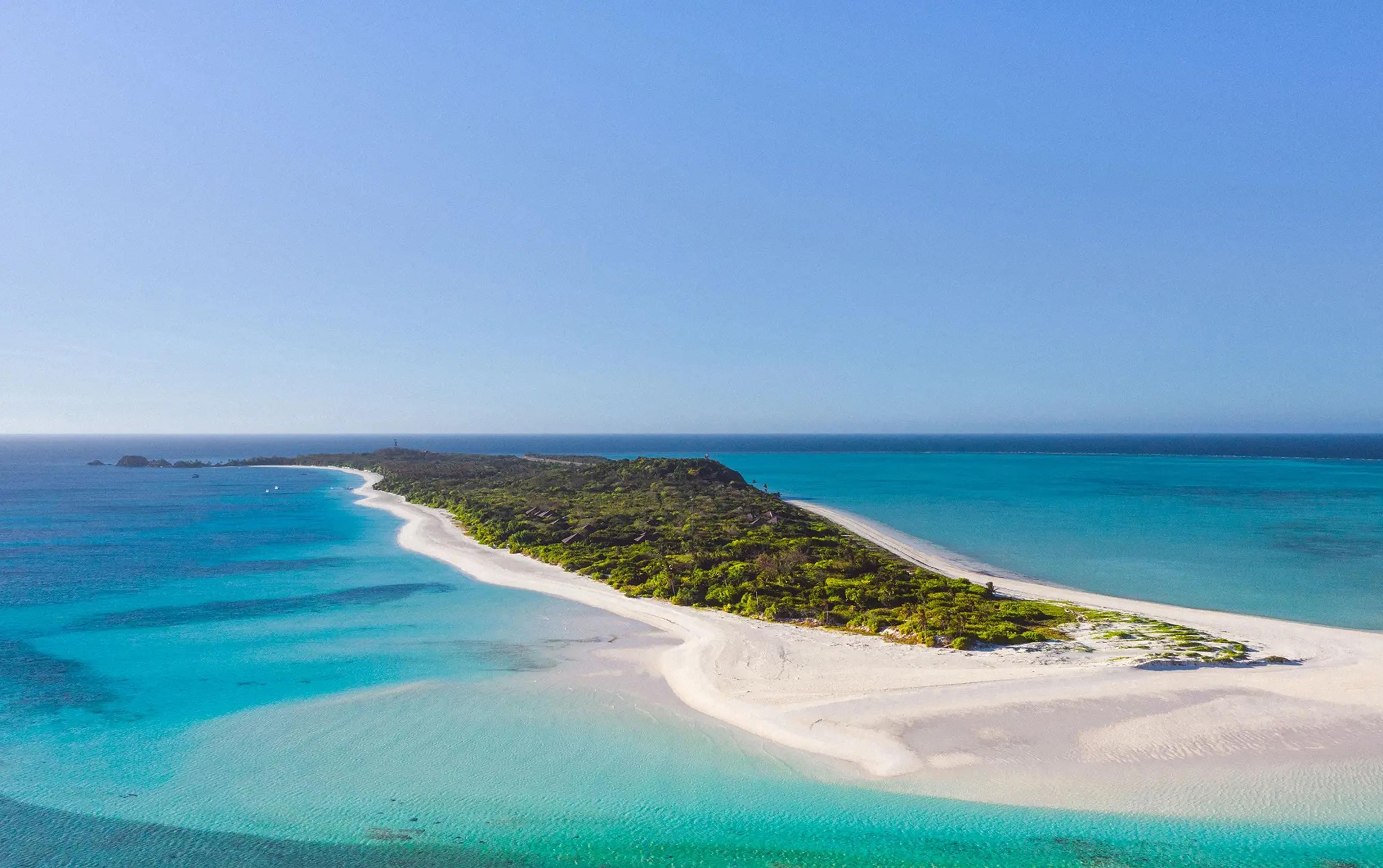 An aerial view of a narrow sandbar covered in green scrub, fringed by pale shallows and deep turquoise sea.