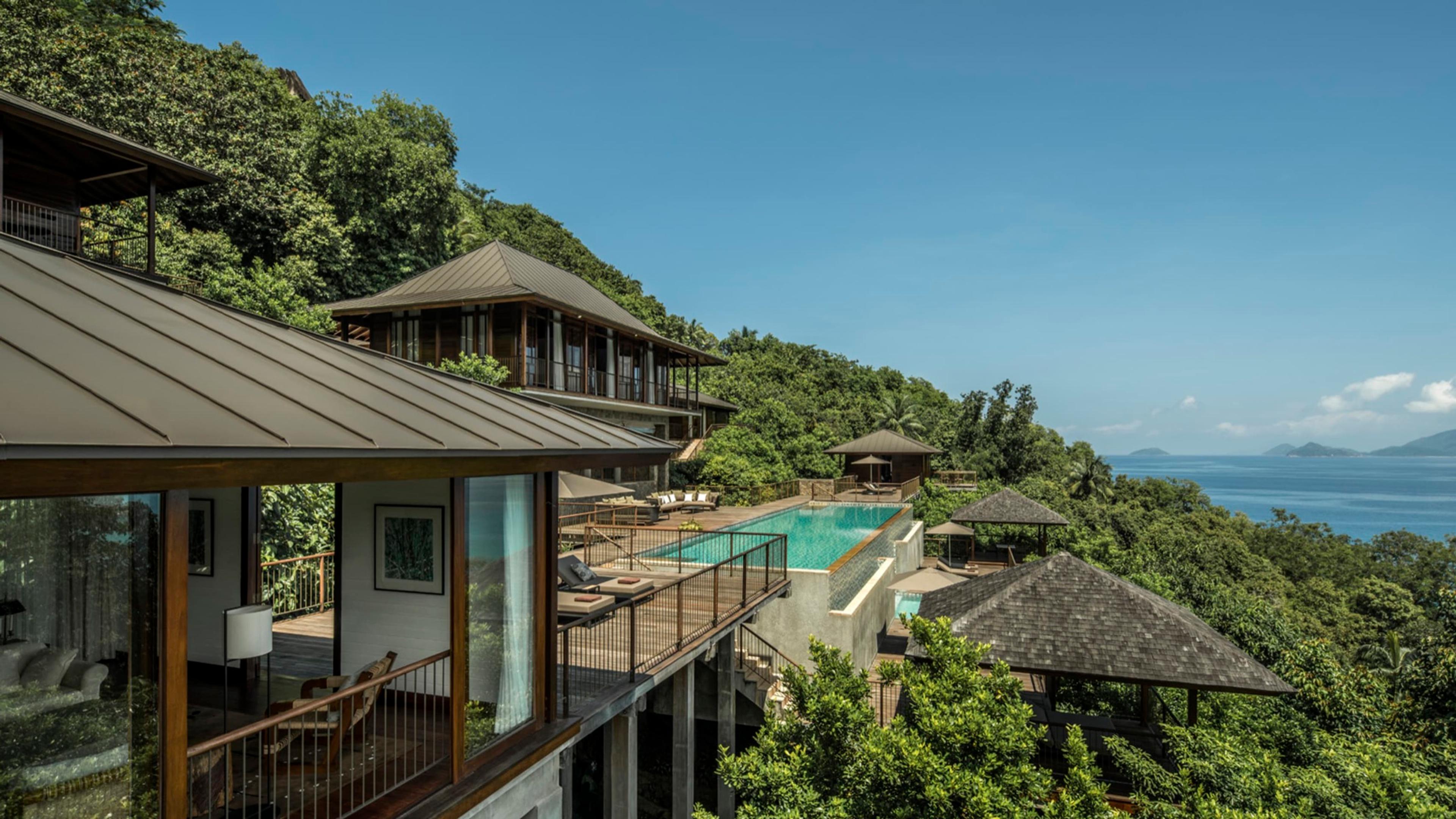 An aerial view of pitched pavilion roofs stepping down a forested hillside to a pool, with the sea beyond.