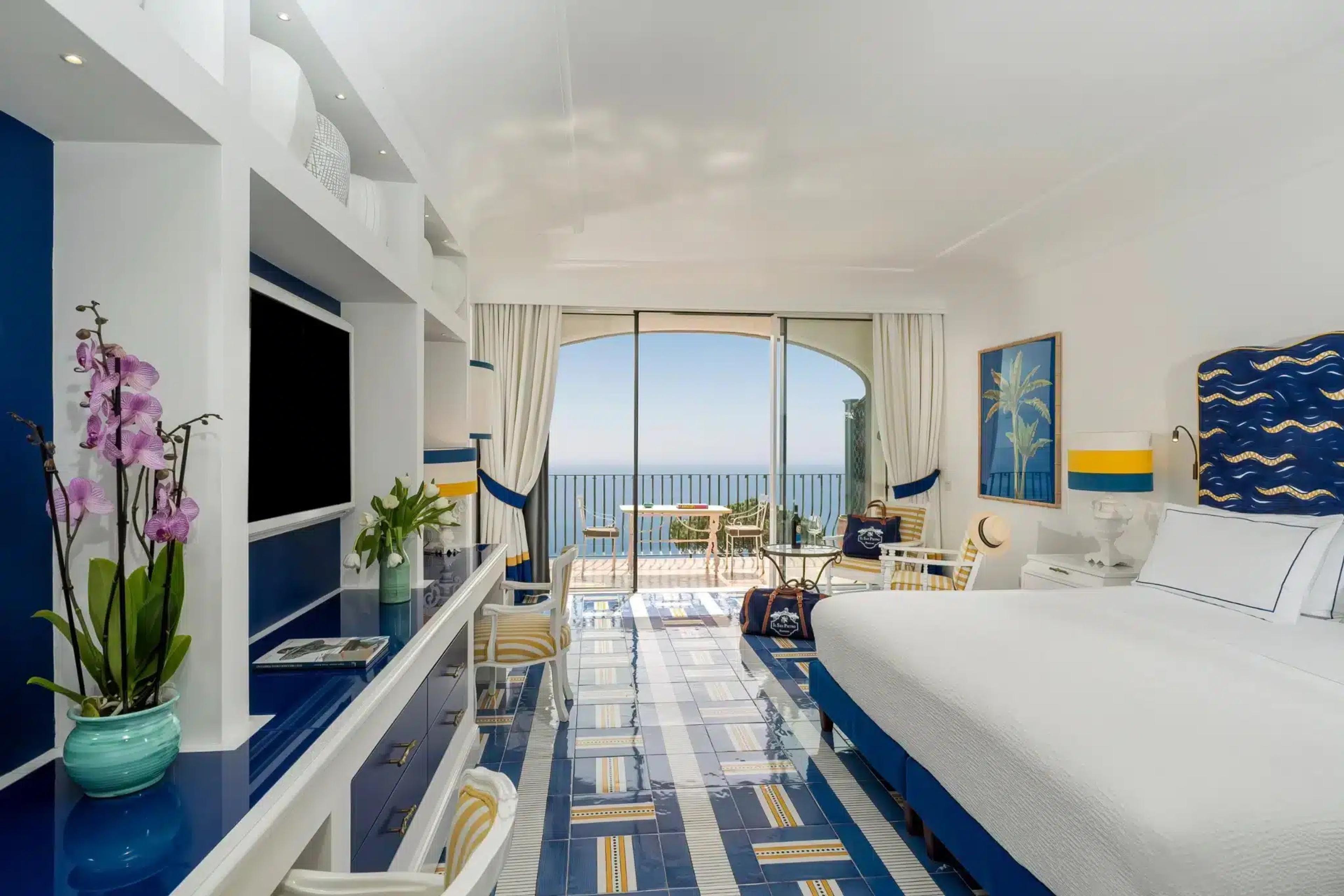 A hotel bedroom in blue and white with orchids on a sideboard, a television, and sliding doors open to a sea-view balcony with a table and chairs.