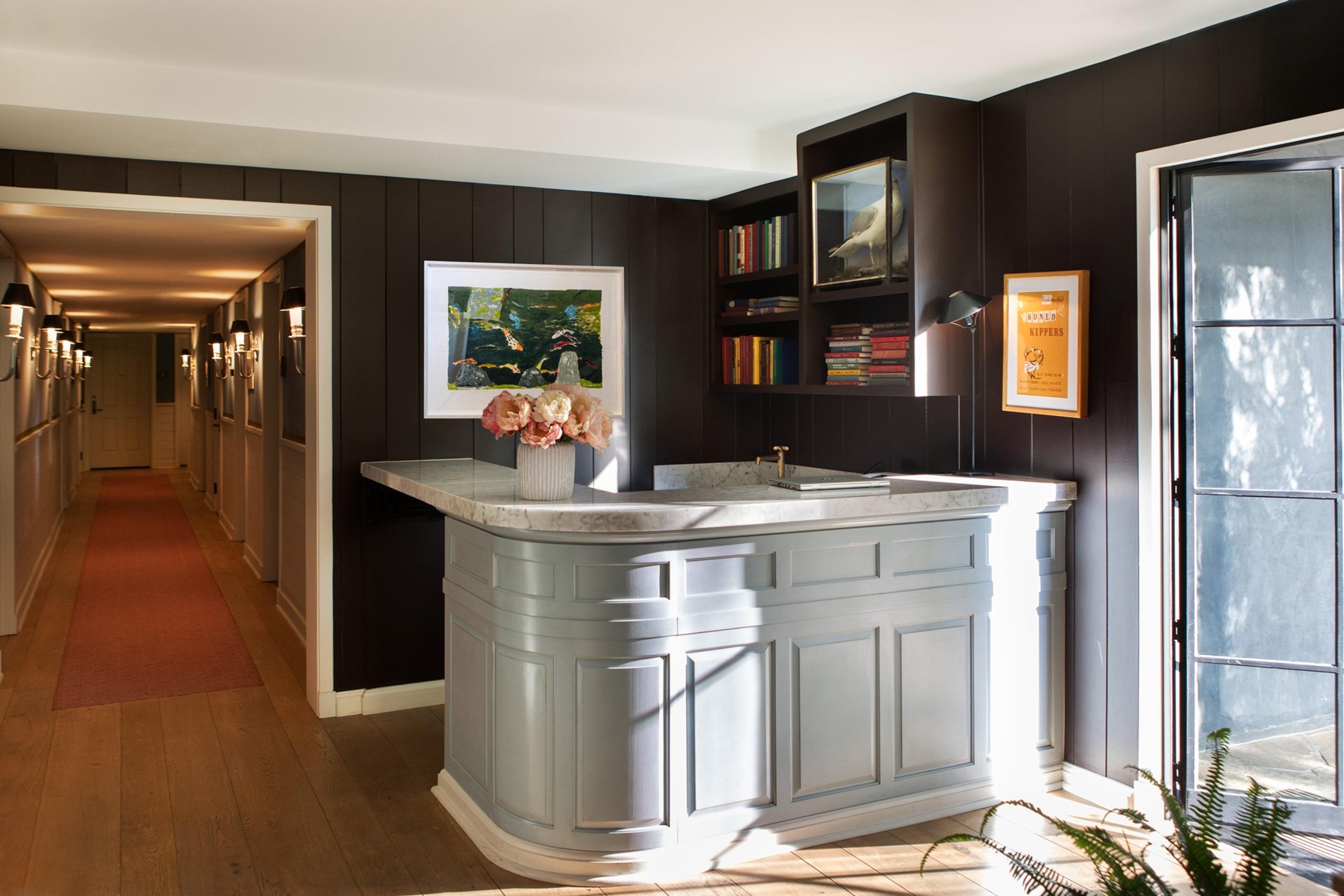 A reception desk with a pale blue marble top, a wall of bookshelves holding a taxidermy bird, and a corridor with wall sconces and a red runner leading off to one side.
