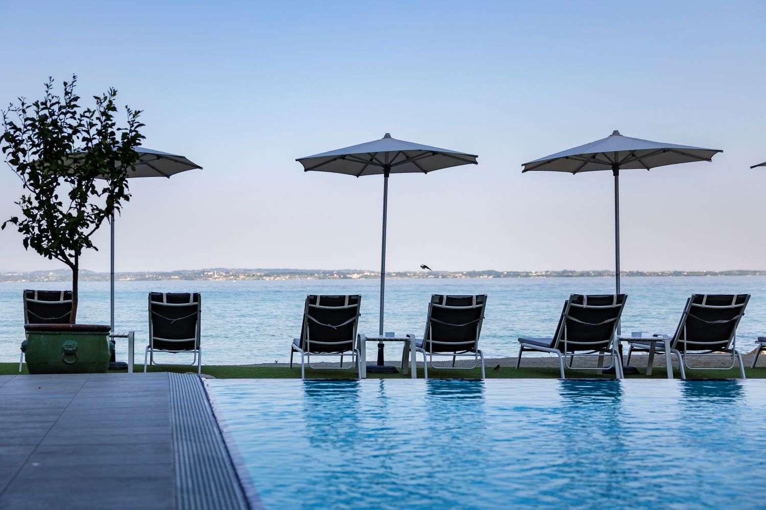 Empty sun loungers and umbrellas at dusk facing the lake, with a small tree in a planter beside the pool's edge.