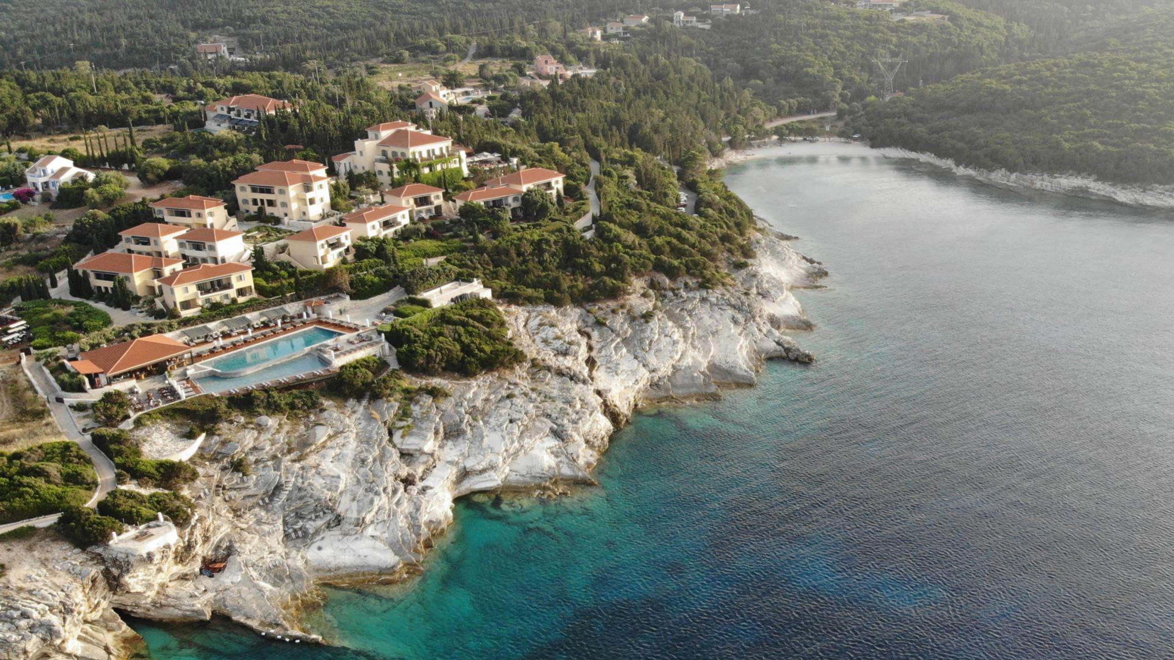 An aerial view of the resort's terracotta-roofed buildings and pool terrace on a pine-covered headland above a turquoise bay.