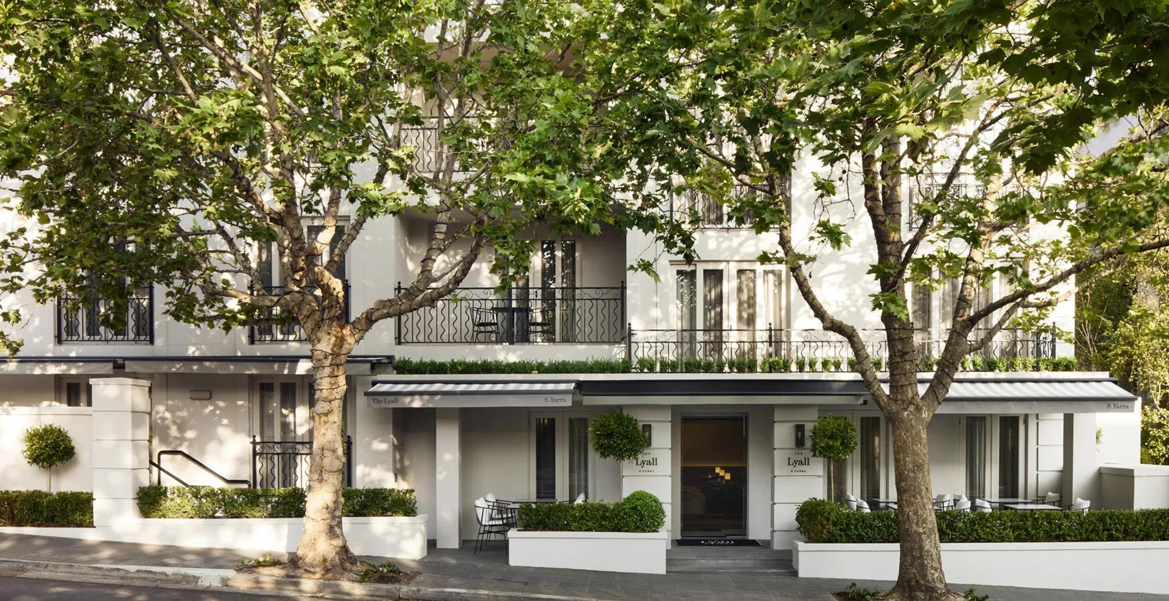 The hotel's white three storey facade seen through two street trees, with wrought iron balconies, an awning over the entrance and outdoor seating below.
