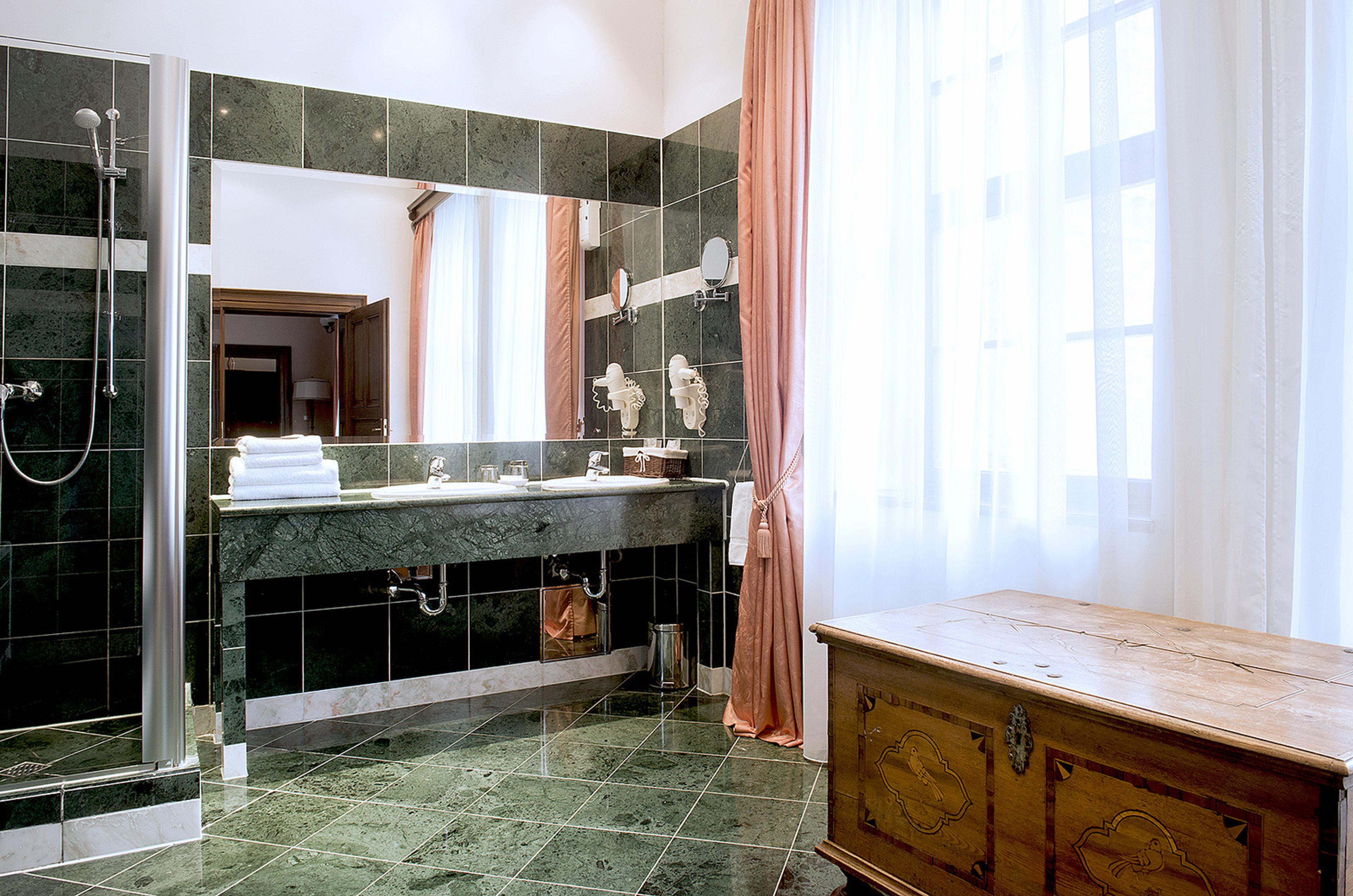 A dark green marble bathroom with two basins, a curtained window and a wooden chest.
