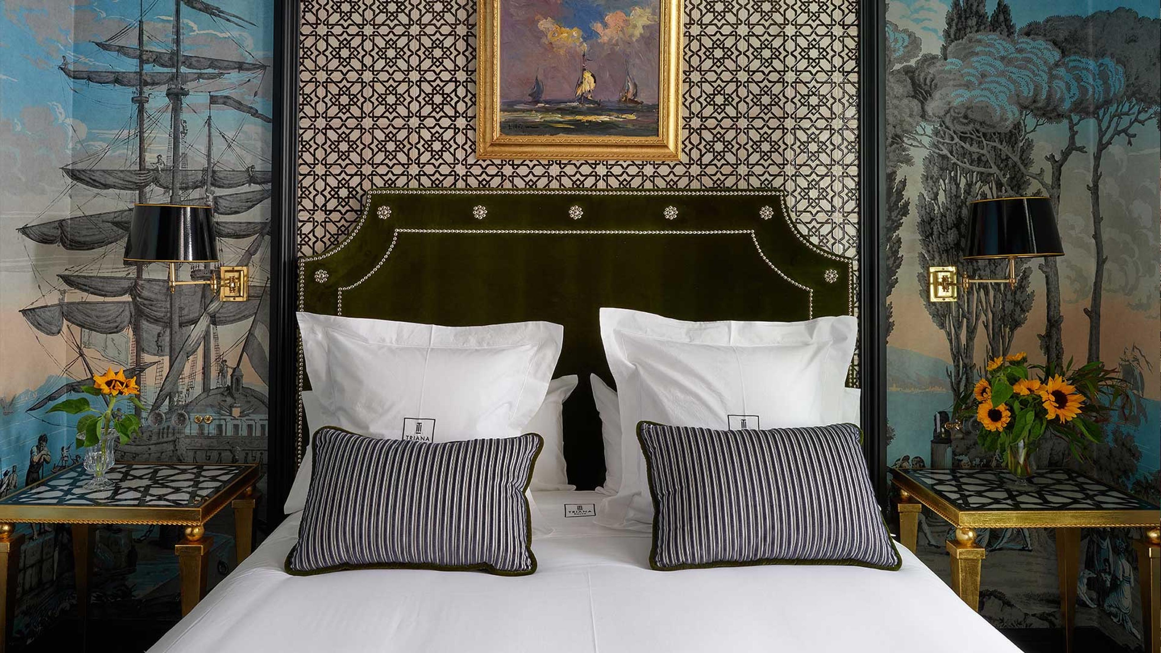A made bed with a green velvet headboard and striped cushions, set against wallpaper of a sailing ship and a framed painting of boats at sea.