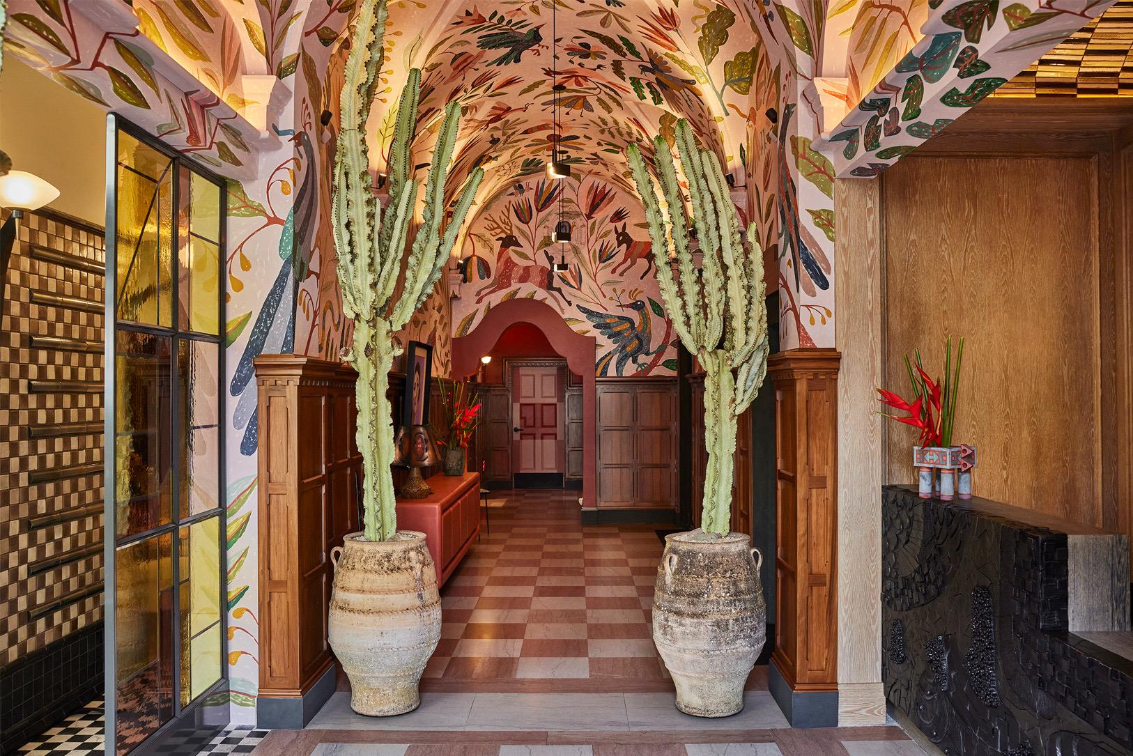 A hotel corridor with a patterned painted ceiling, two large potted cacti, and a black and white checkerboard floor leading toward a lit doorway.