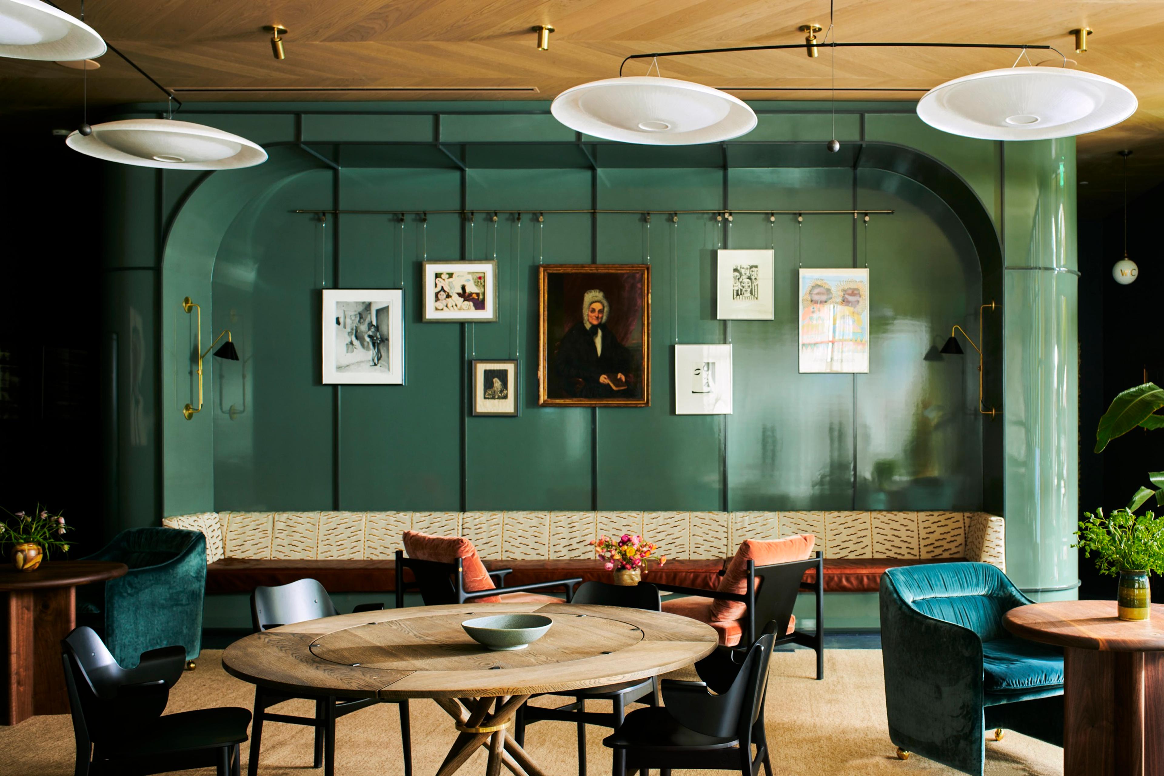 A round wooden table and armchairs in a lounge, with a green lacquered wall behind a long banquette and framed pictures.