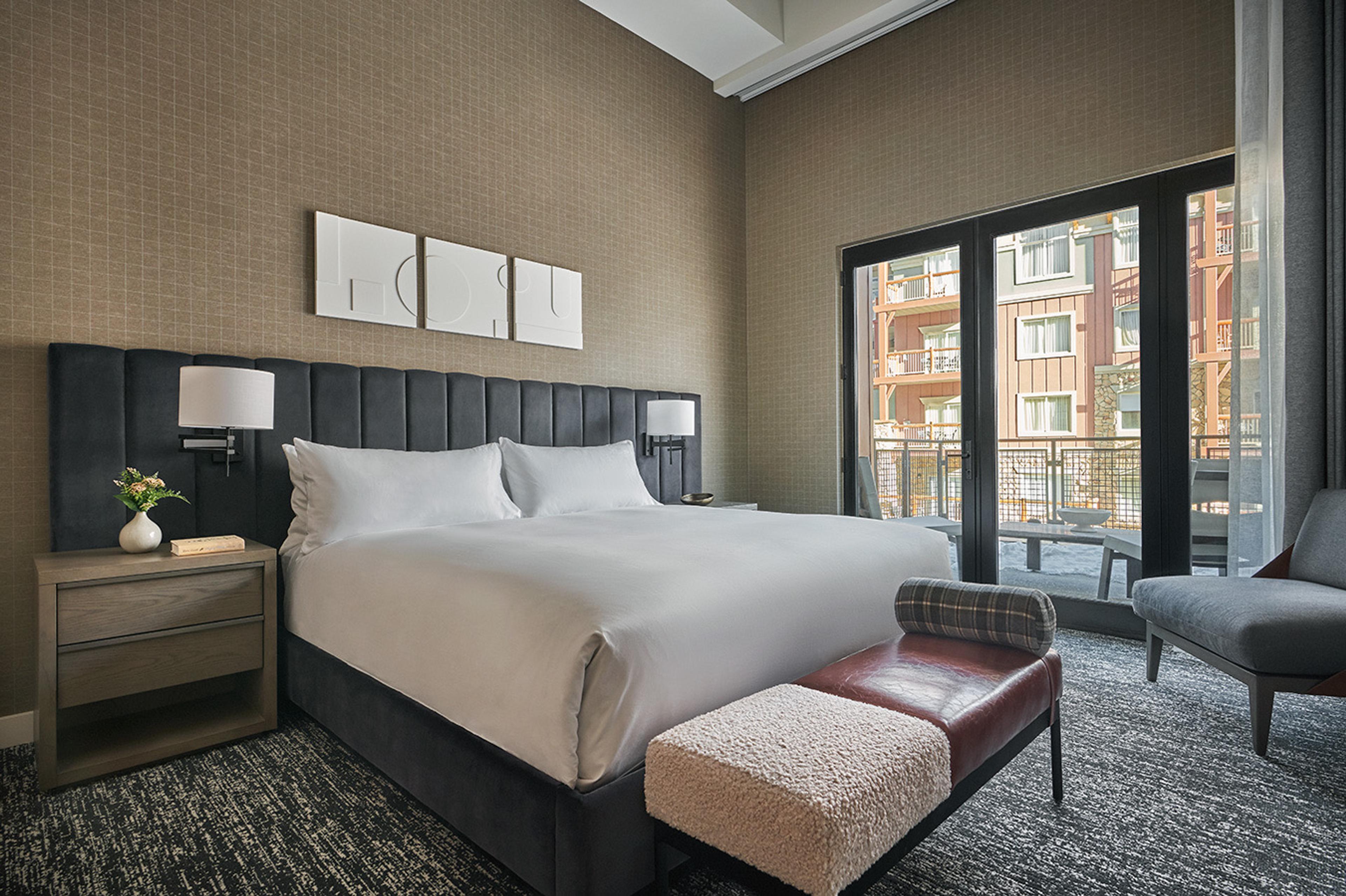 A king bed with white bedding and a tall grey upholstered headboard, a bench at its foot, and a glass door onto a balcony that looks across to another building.