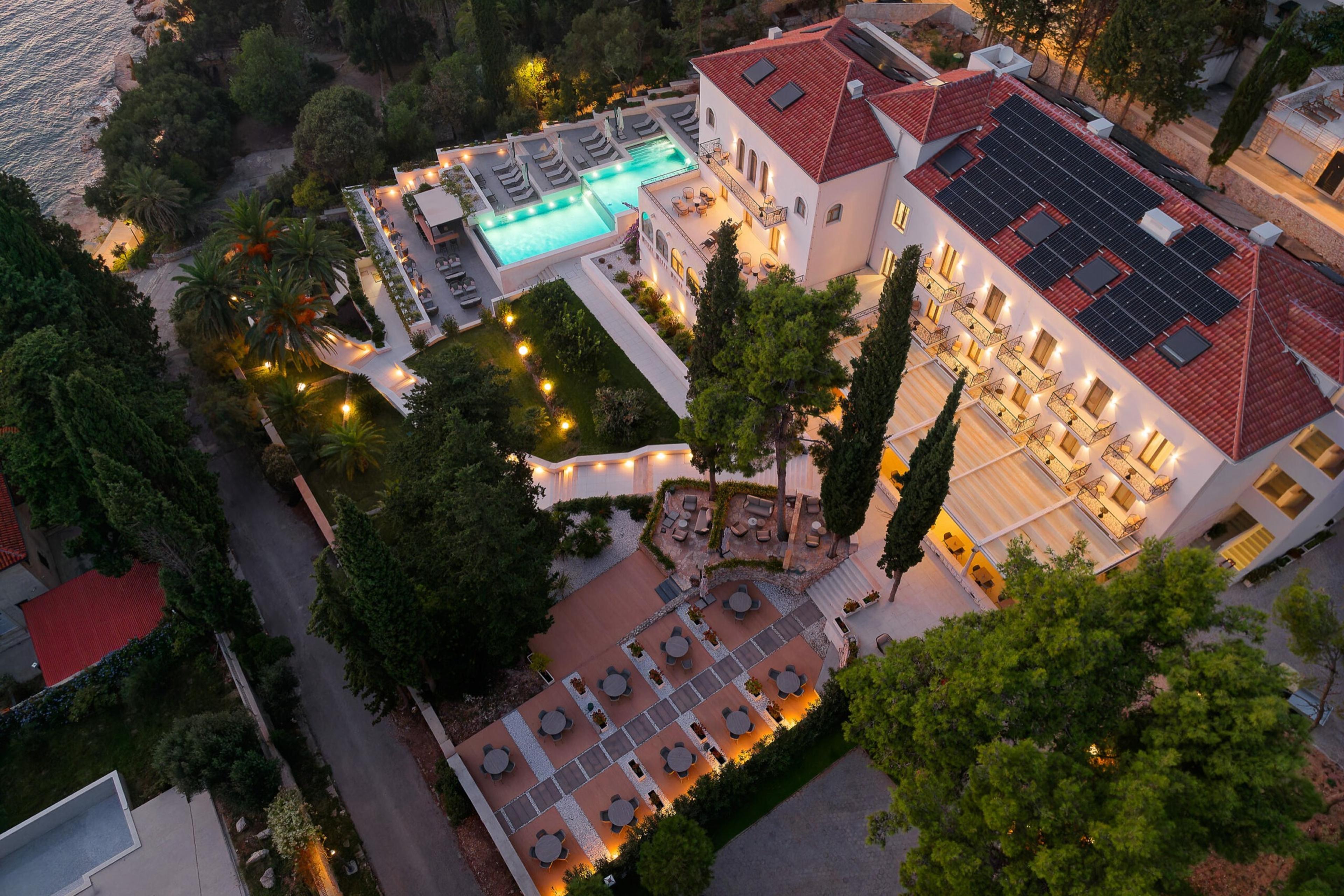 An aerial view at dusk of a hotel building with a red tiled roof, a lit swimming pool, and a restaurant terrace set with round tables, surrounded by trees and the sea.