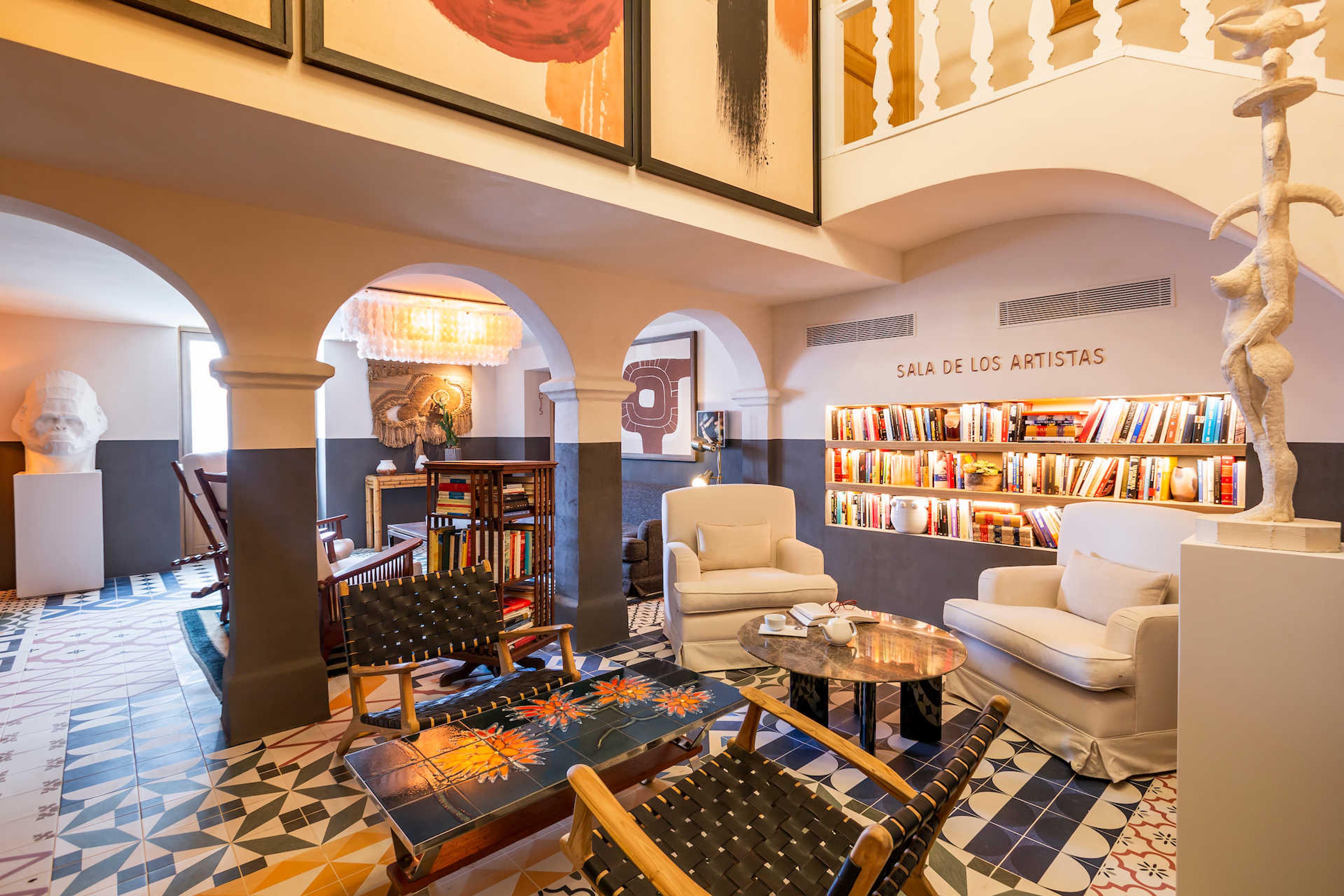 A lounge with arched openings, a cream sofa and armchairs, shelves of books lit from behind, and a white bust on a plinth on a patterned tiled floor.