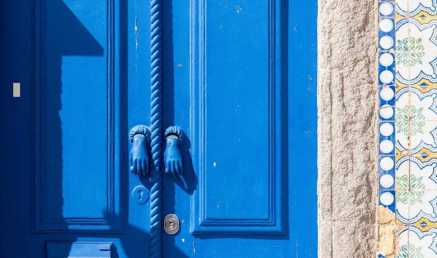 A blue painted double door with hand-shaped brass knockers, beside a wall of patterned blue and yellow tiles.