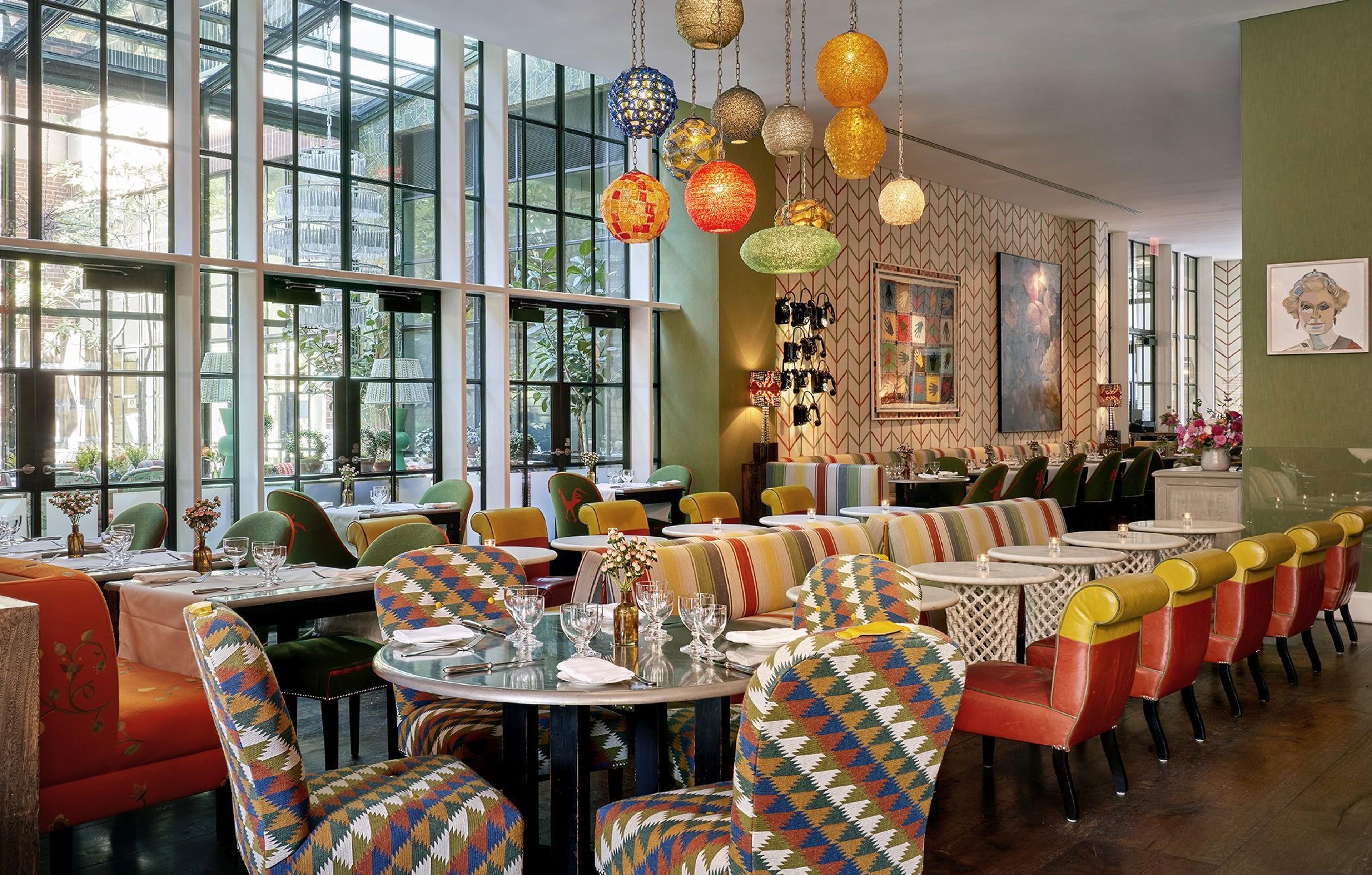 A restaurant dining room with round tables set for service, striped banquette seating, and a cluster of colourful glass lanterns hanging from the ceiling in front of floor to ceiling windows.
