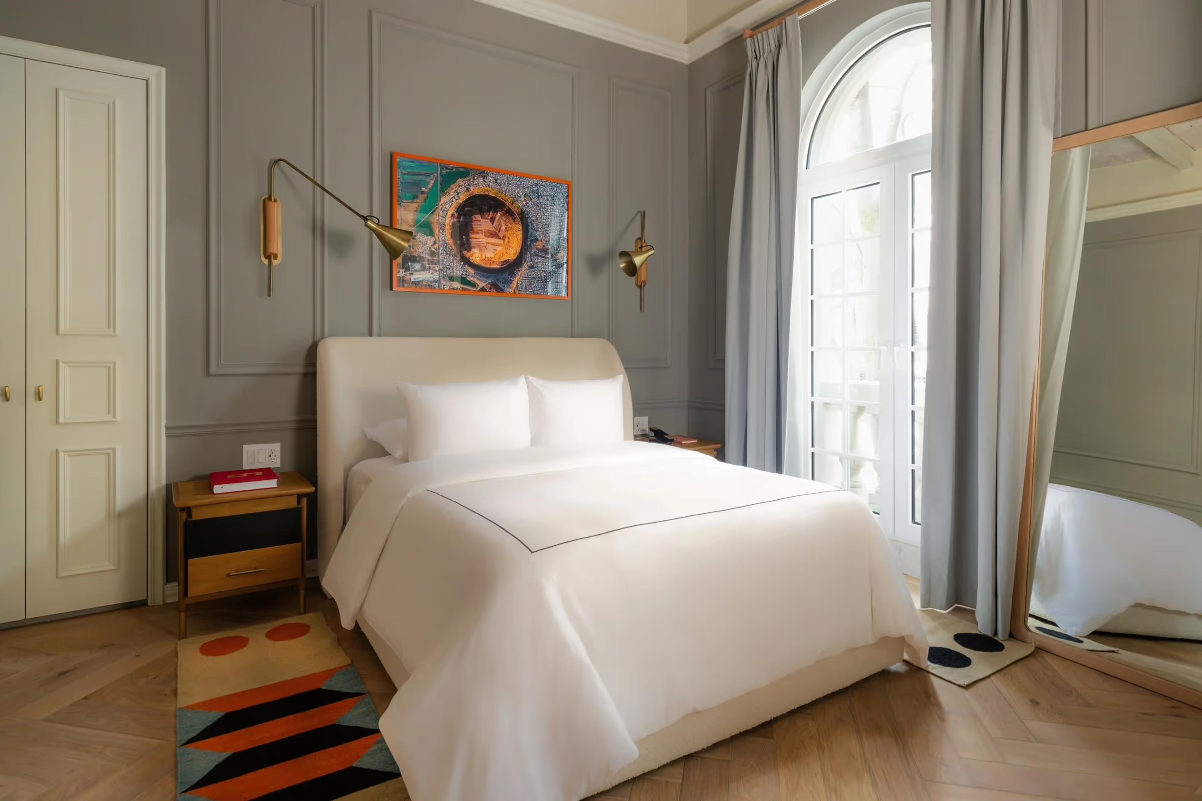 A bedroom with grey panelled walls, a cream upholstered bed, a colourful abstract painting above the headboard, and an arched door open to a balcony.