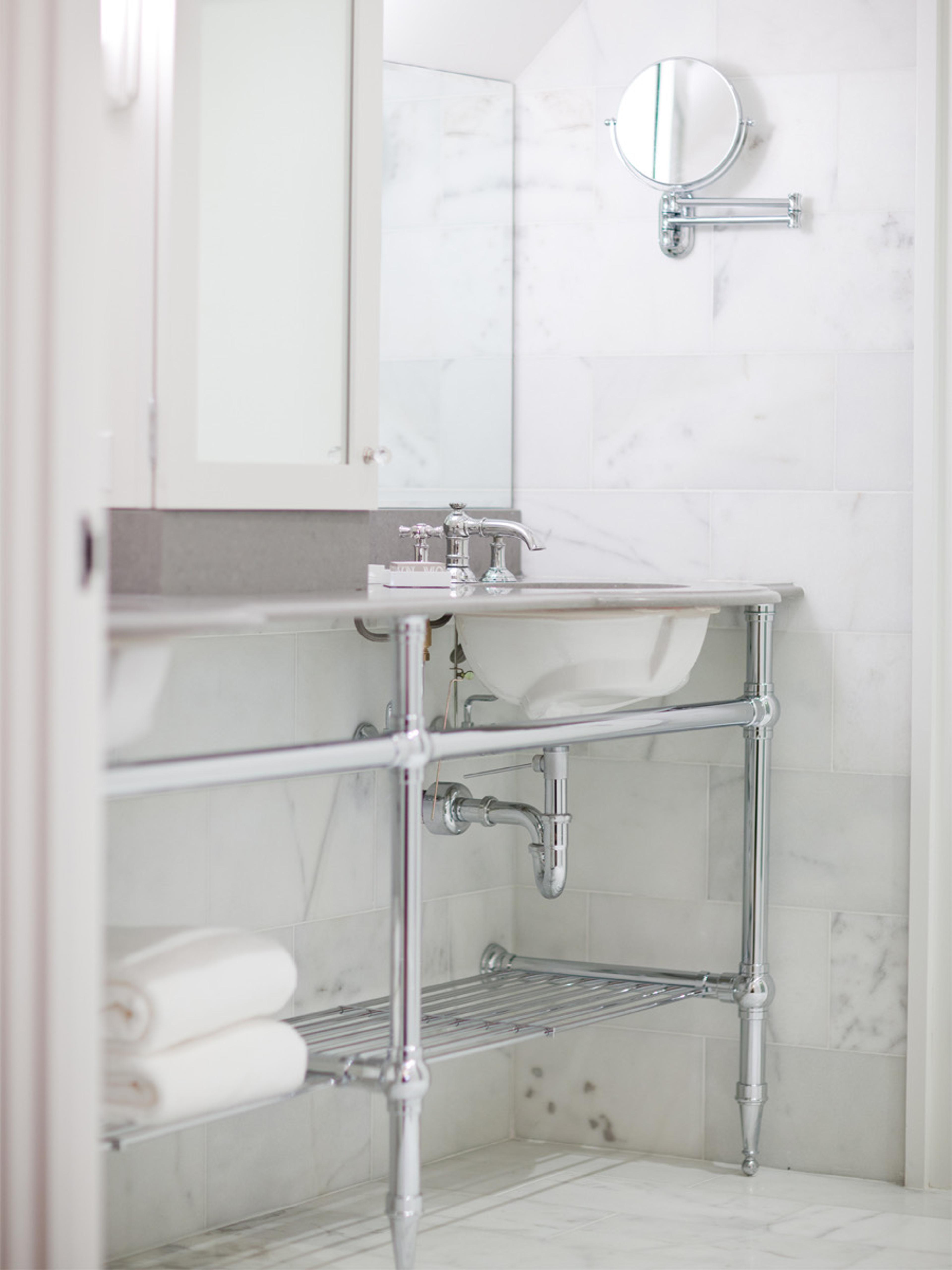 A bathroom with a marble-topped chrome washstand, folded white towels on a lower shelf, and a mirror beside a swing-arm magnifying mirror.
