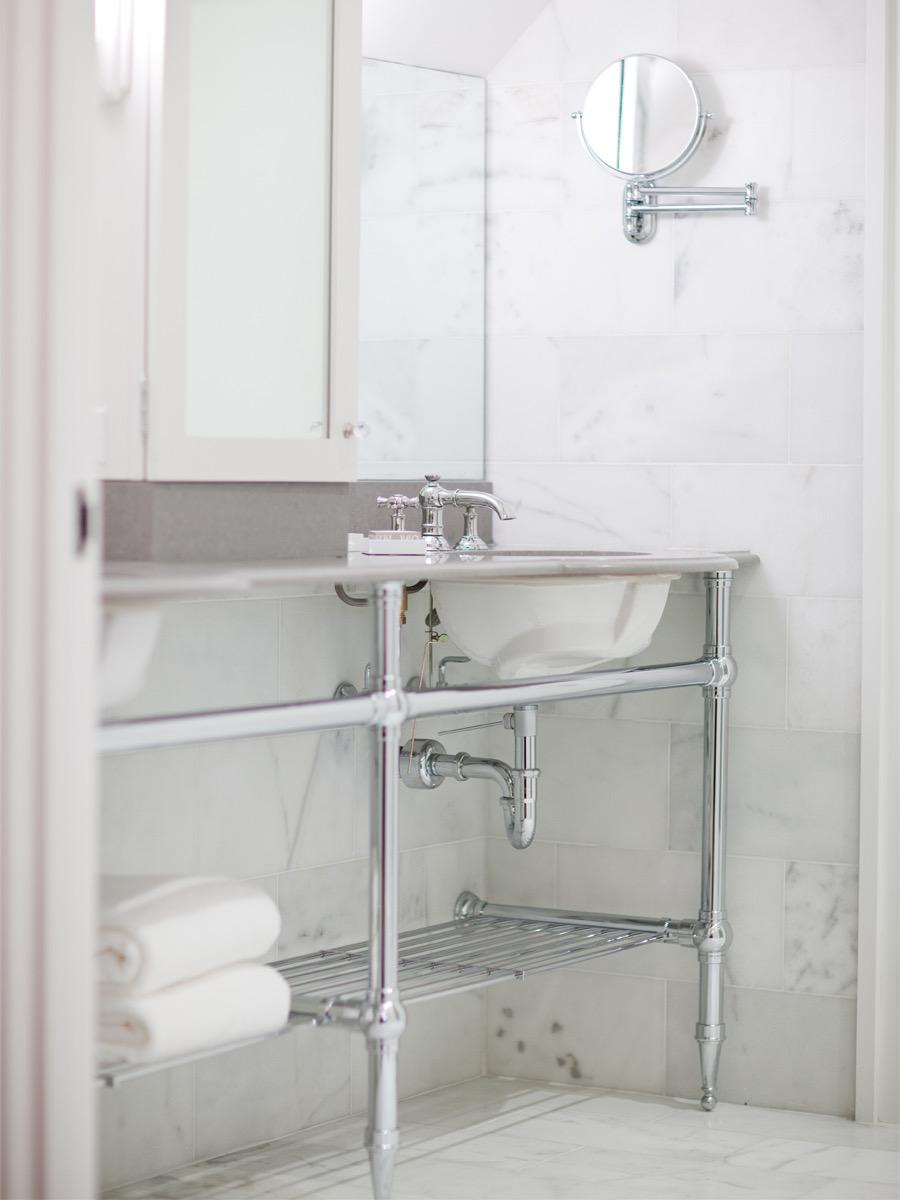 A bathroom with a marble-topped chrome washstand, folded white towels on a lower shelf, and a mirror beside a swing-arm magnifying mirror.