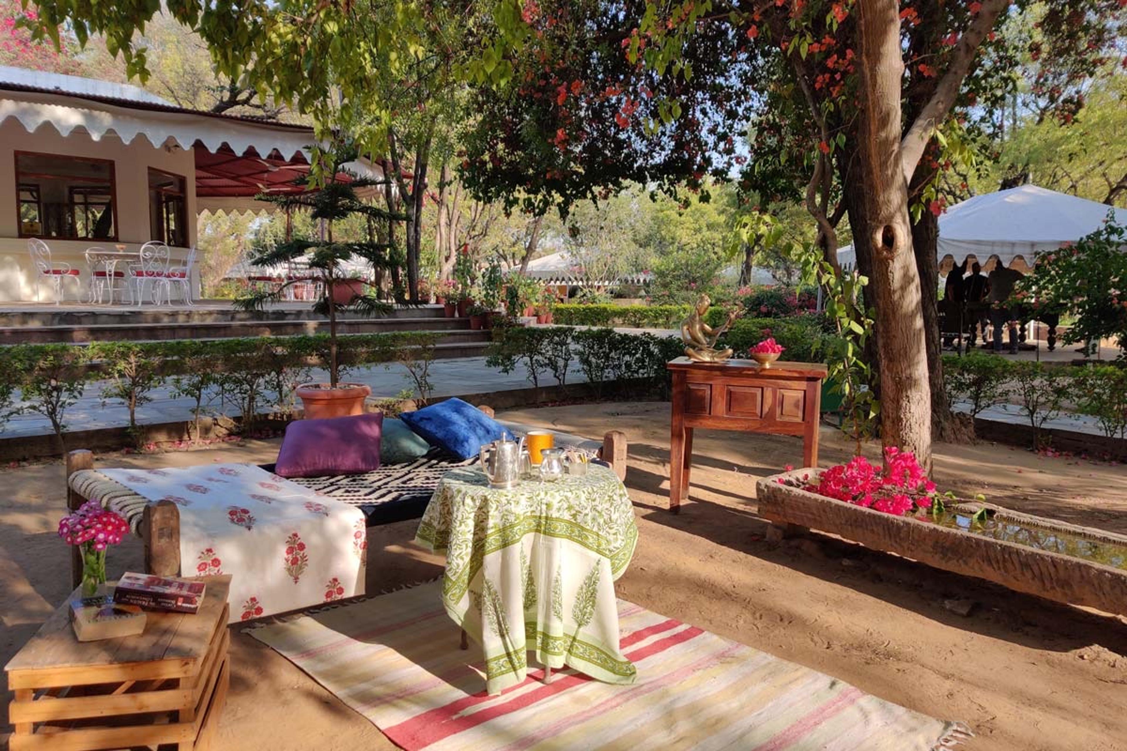 A shaded garden terrace with a cushioned day bed, a round table laid for tea, and a bench of trailing pink flowers, with a marquee visible beyond the trees.
