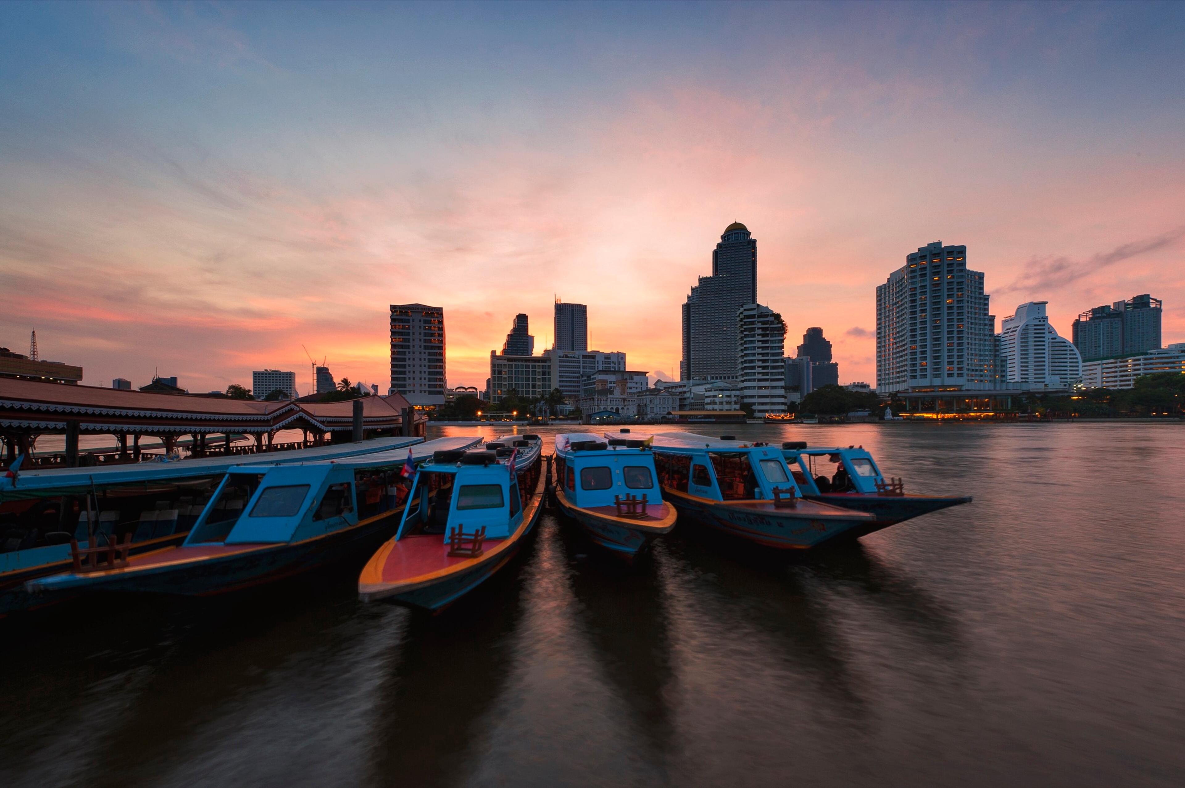 The hotel's teak boats moored on the Chao Phraya at dusk, with the city lit behind
