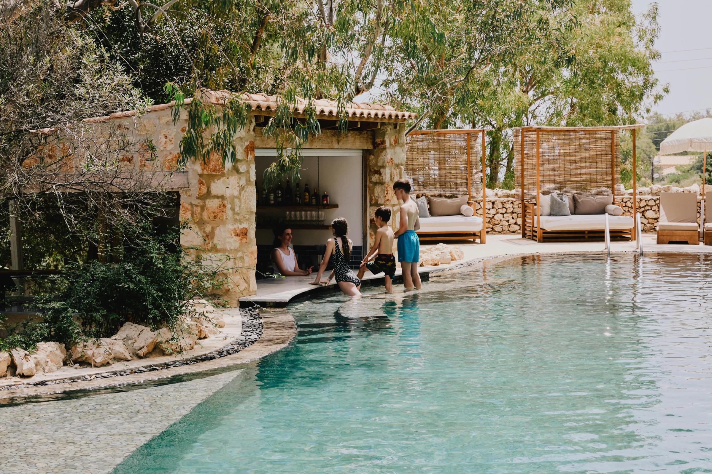 A stone poolside building with a bar inside, people wading at its edge, and wicker daybeds beside an outdoor pool.