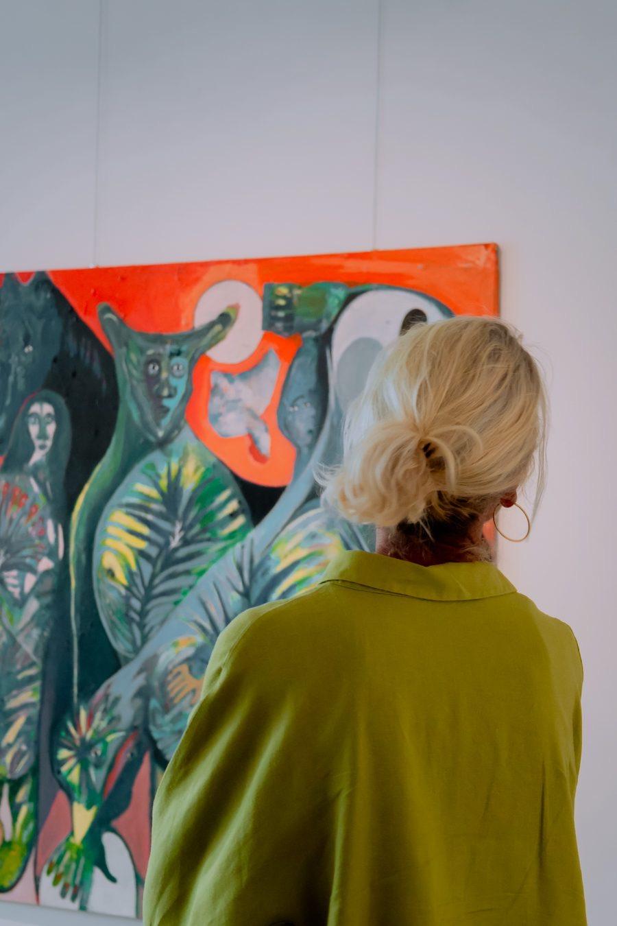 A woman seen from behind, with her hair in a bun and a green jacket, looks at a large painting of green and orange figures on a white wall.
