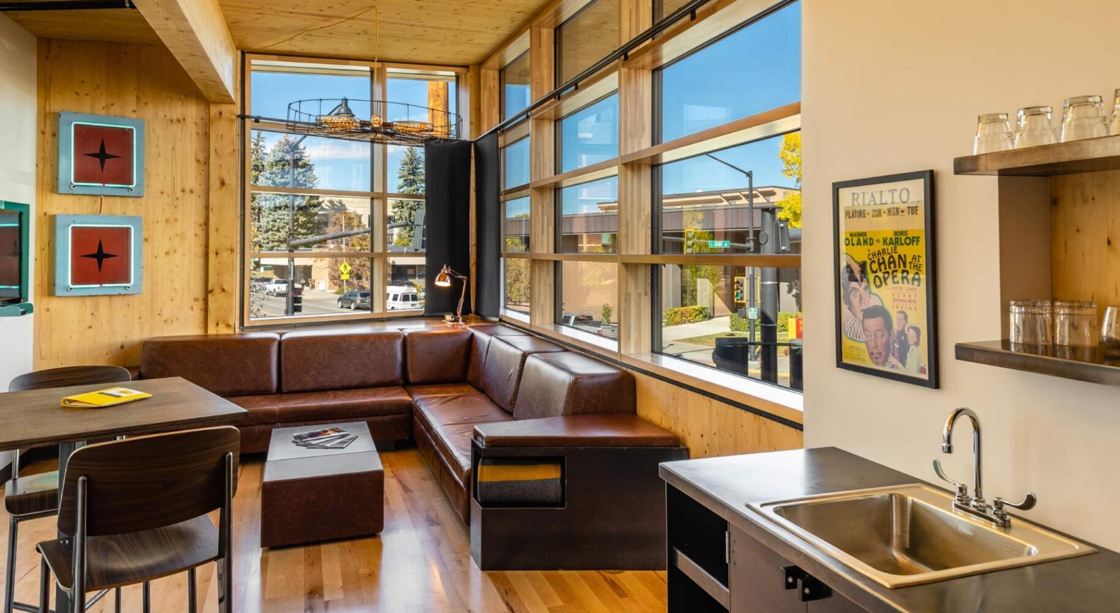 A sitting area with a brown leather banquette along a wall of tall windows onto a street, with a dark kitchenette sink in the foreground and framed posters on the wall.