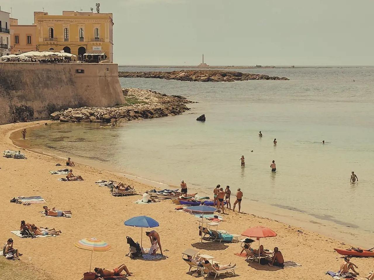An elevated view of a sandy beach beneath old stone city walls, with sunbathers, umbrellas and swimmers in clear turquoise water, and a yellow building on the headland above.