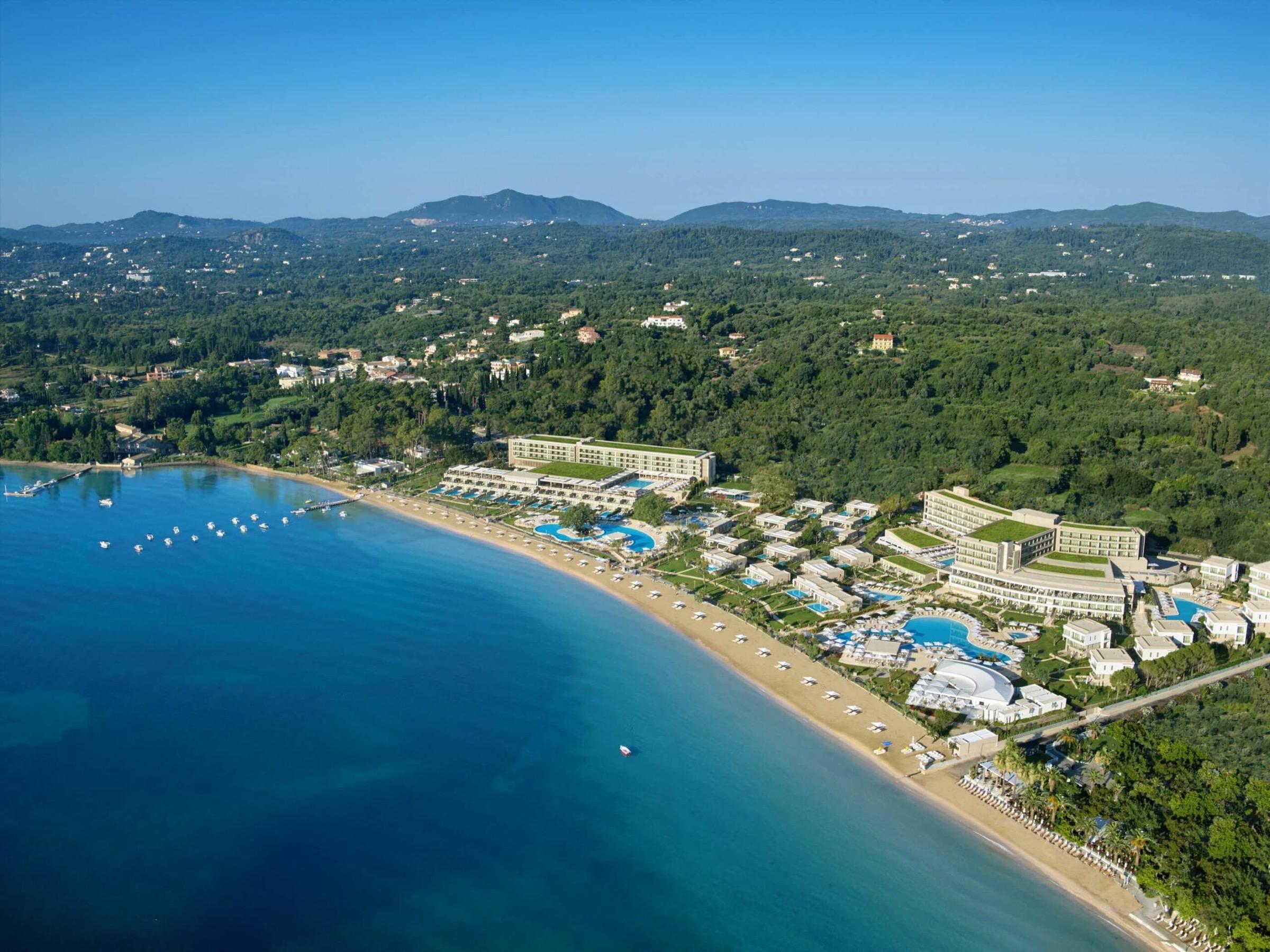 An aerial view of a beachfront hotel complex with several buildings, pools and a long sandy beach, backed by hills and the sea beyond.
