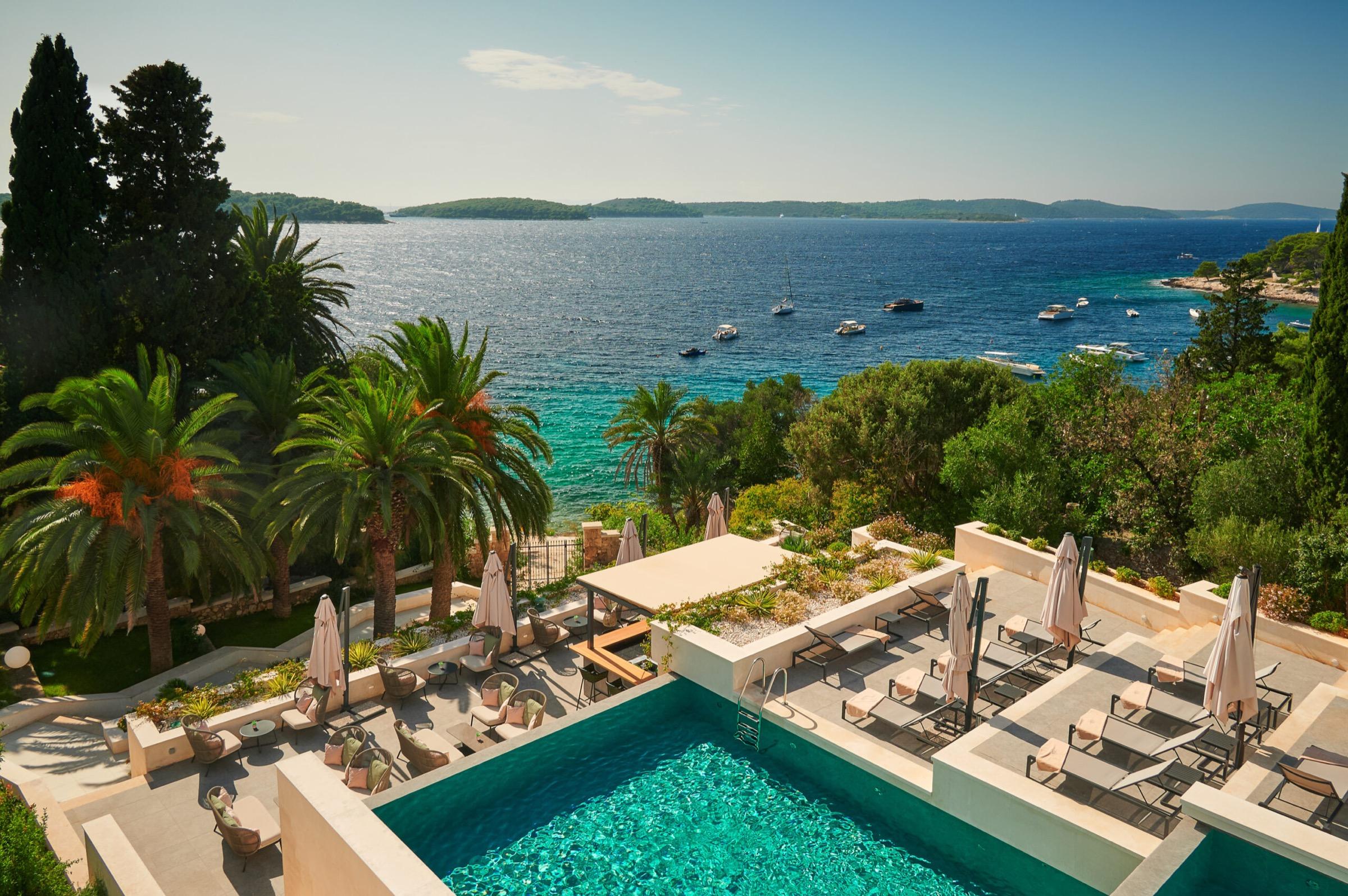 A swimming pool terrace in daylight with the sea and a scattering of islands in the distance, sun loungers lined up along one edge.