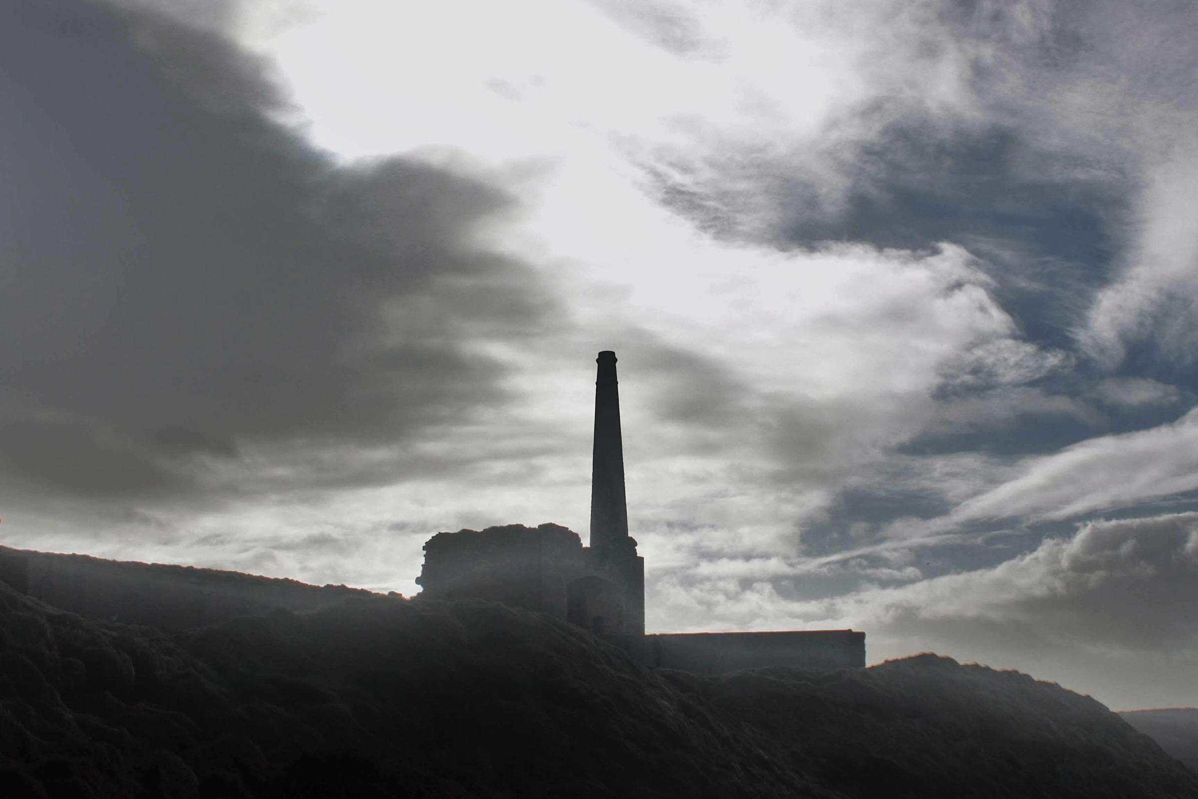The engine house of an old tin mine on a ridge, black against a grey sky.