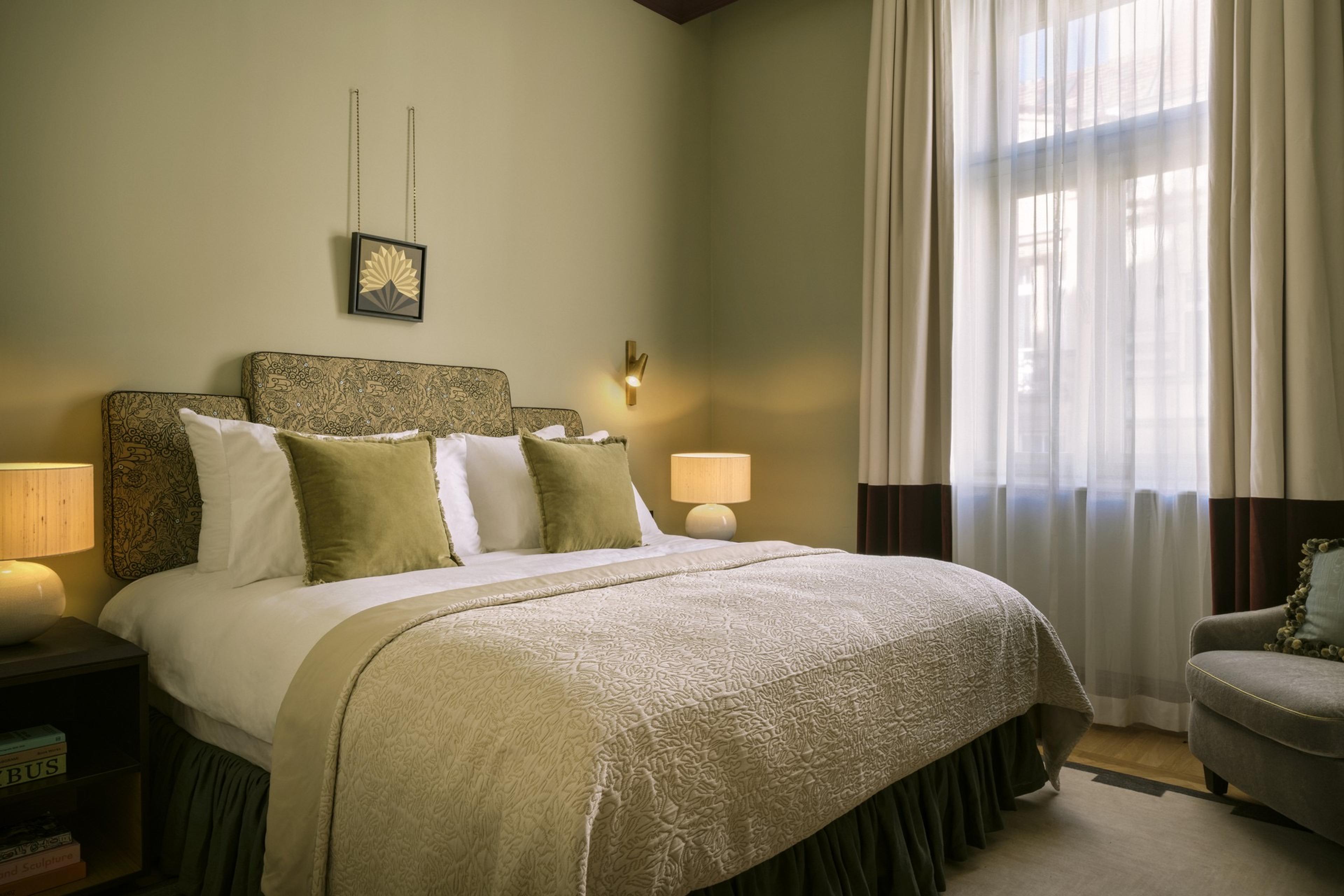 A bedroom with a patterned fabric headboard, white bedding with sage green cushions, two bedside lamps, and a curtained window showing nearby rooftops.