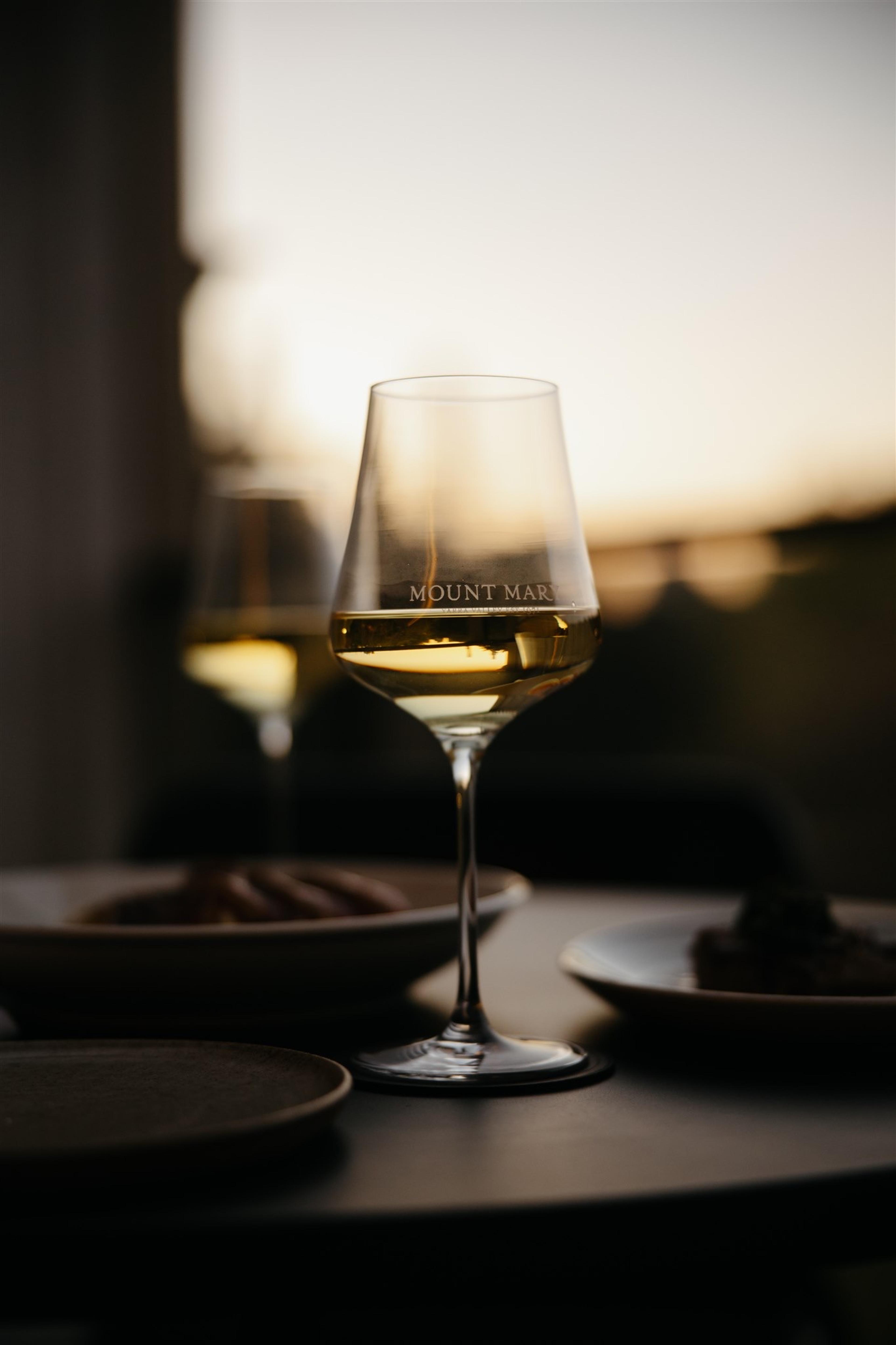 two wine glasses and plates of food on a table outdoors at dusk, with trees blurred in the background