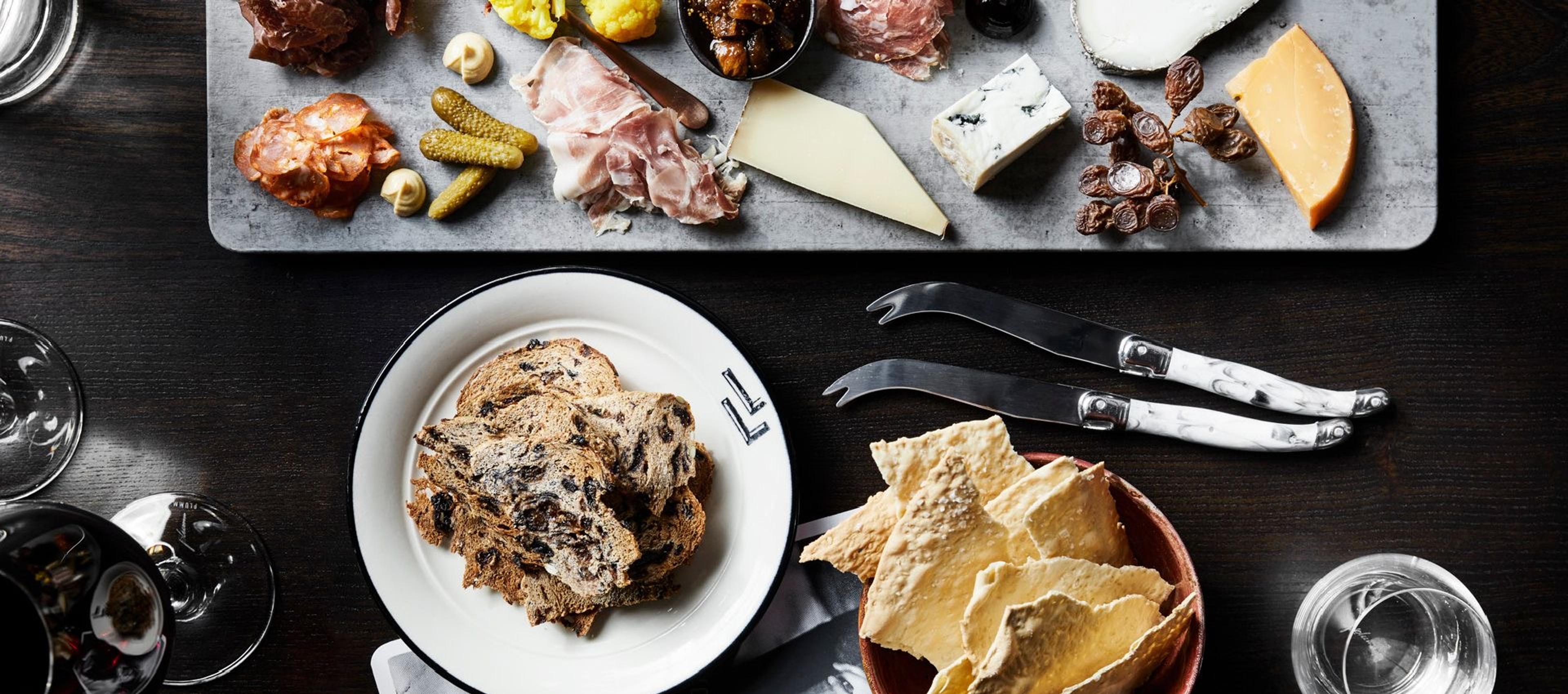 an overhead view of a charcuterie and cheese board with crackers, bread and a glass of wine on a dark wood table