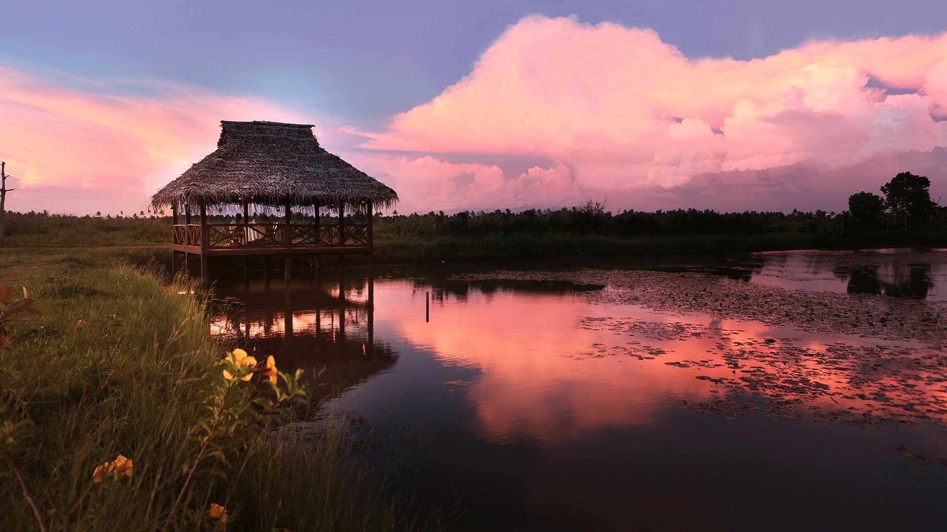 A thatched gazebo over a lake at sunset, under a sky of pink clouds
