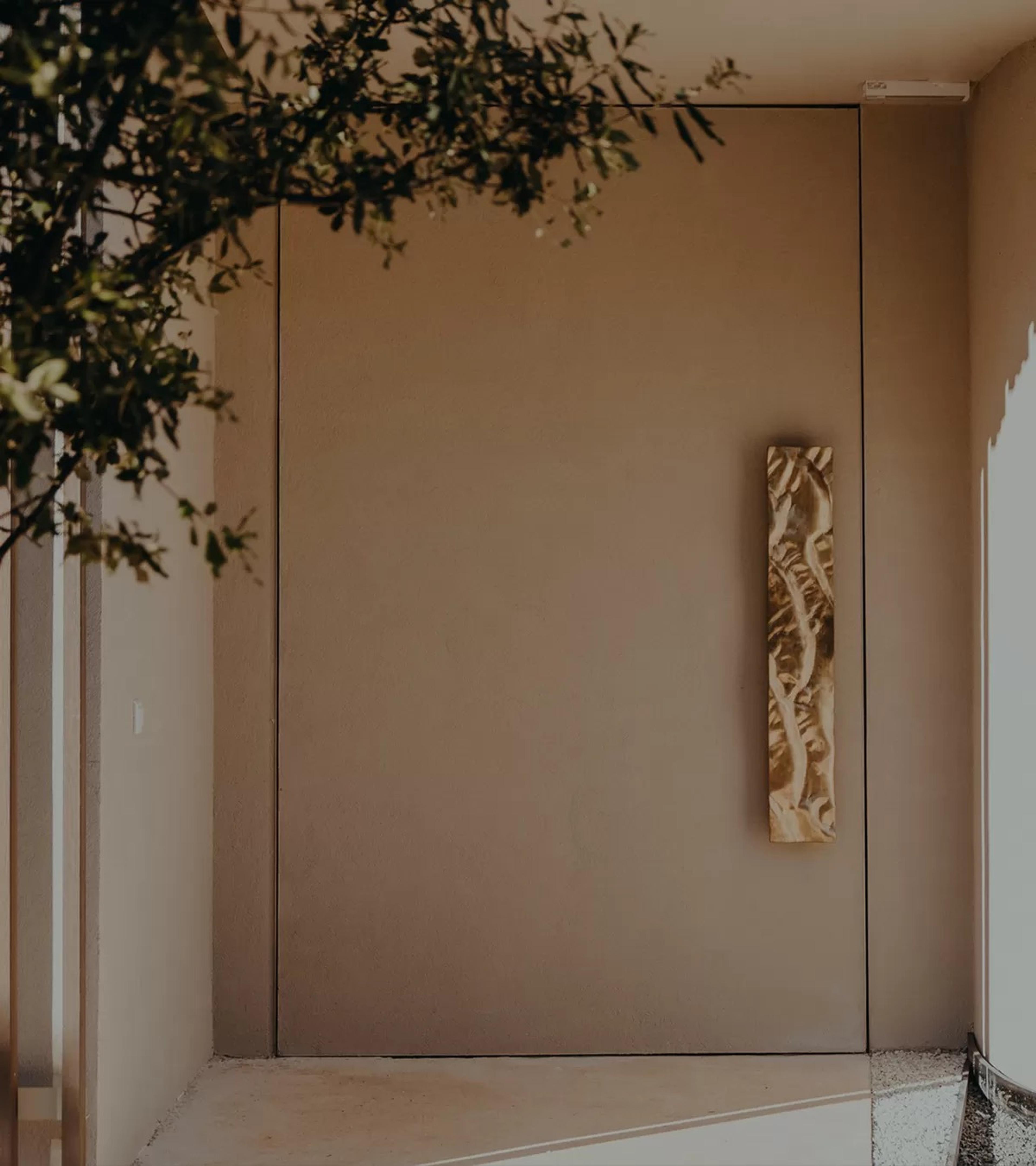 A pale wall with a bronze relief panel beside a recessed doorway, and an olive branch overhead.