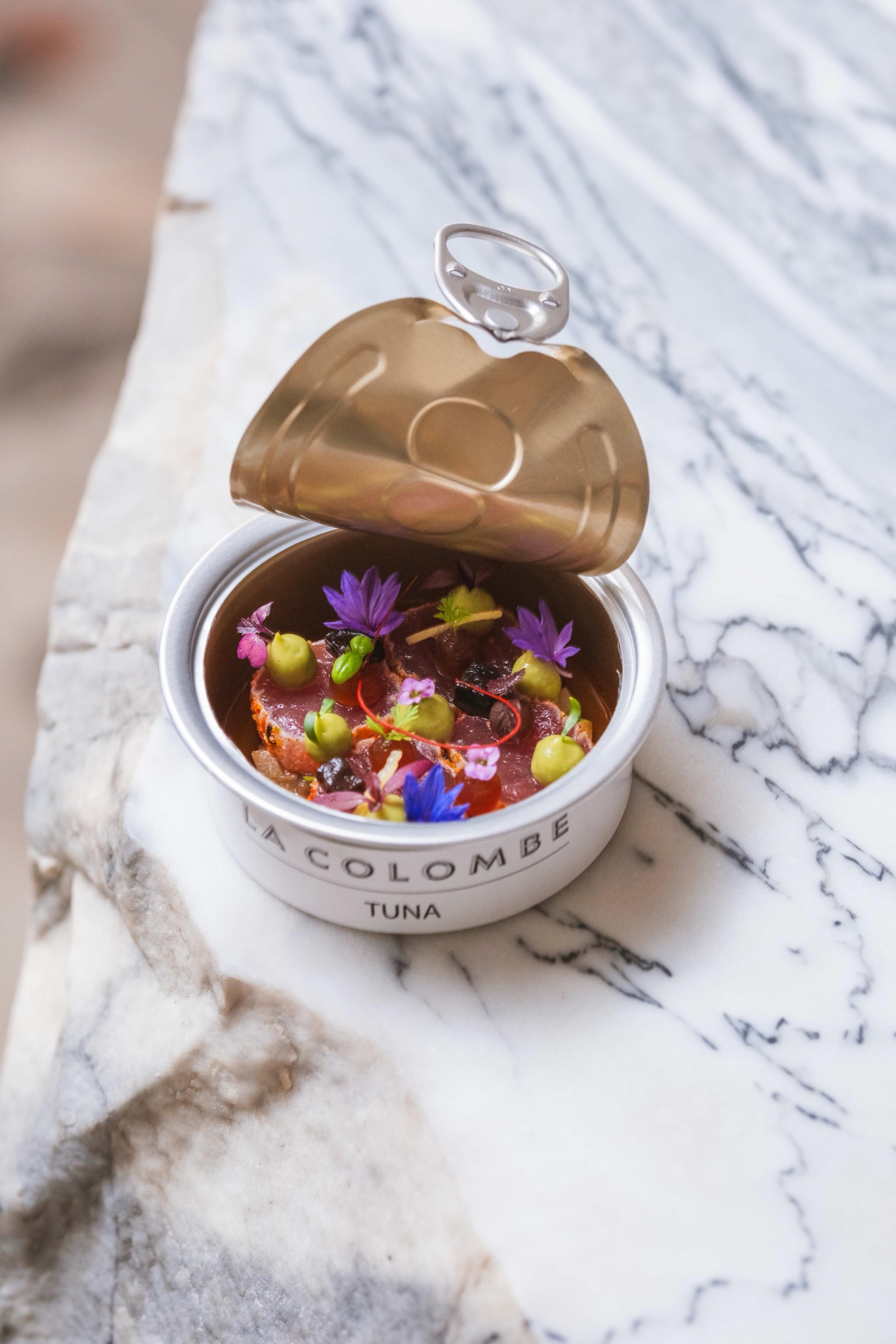 An opened tin labelled La Colombe on a marble table, filled with tuna and edible flowers
