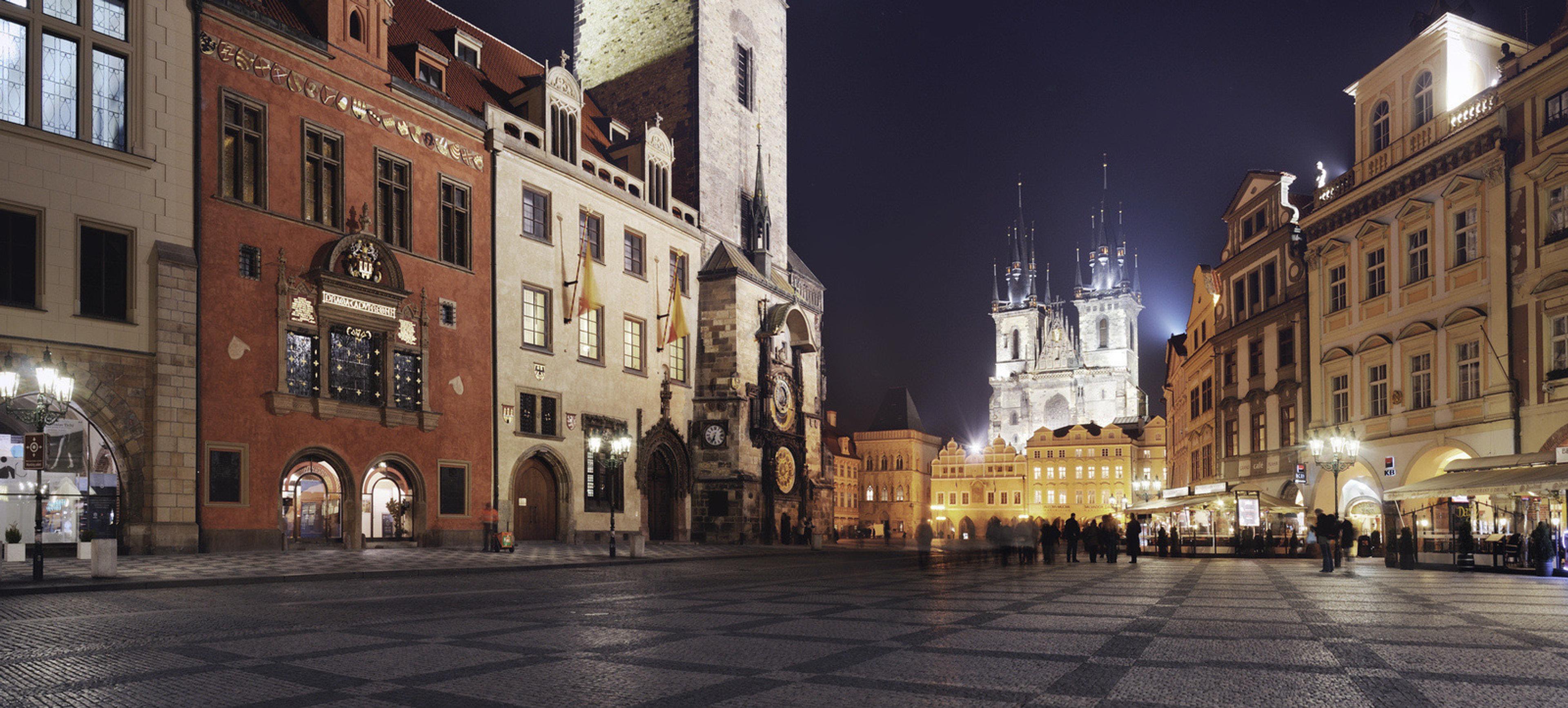 A cobbled square at night, with floodlit historic buildings and the twin spires of a gothic church in the distance.