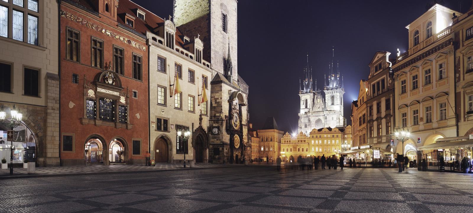 A cobbled square at night, with floodlit historic buildings and the twin spires of a gothic church in the distance.