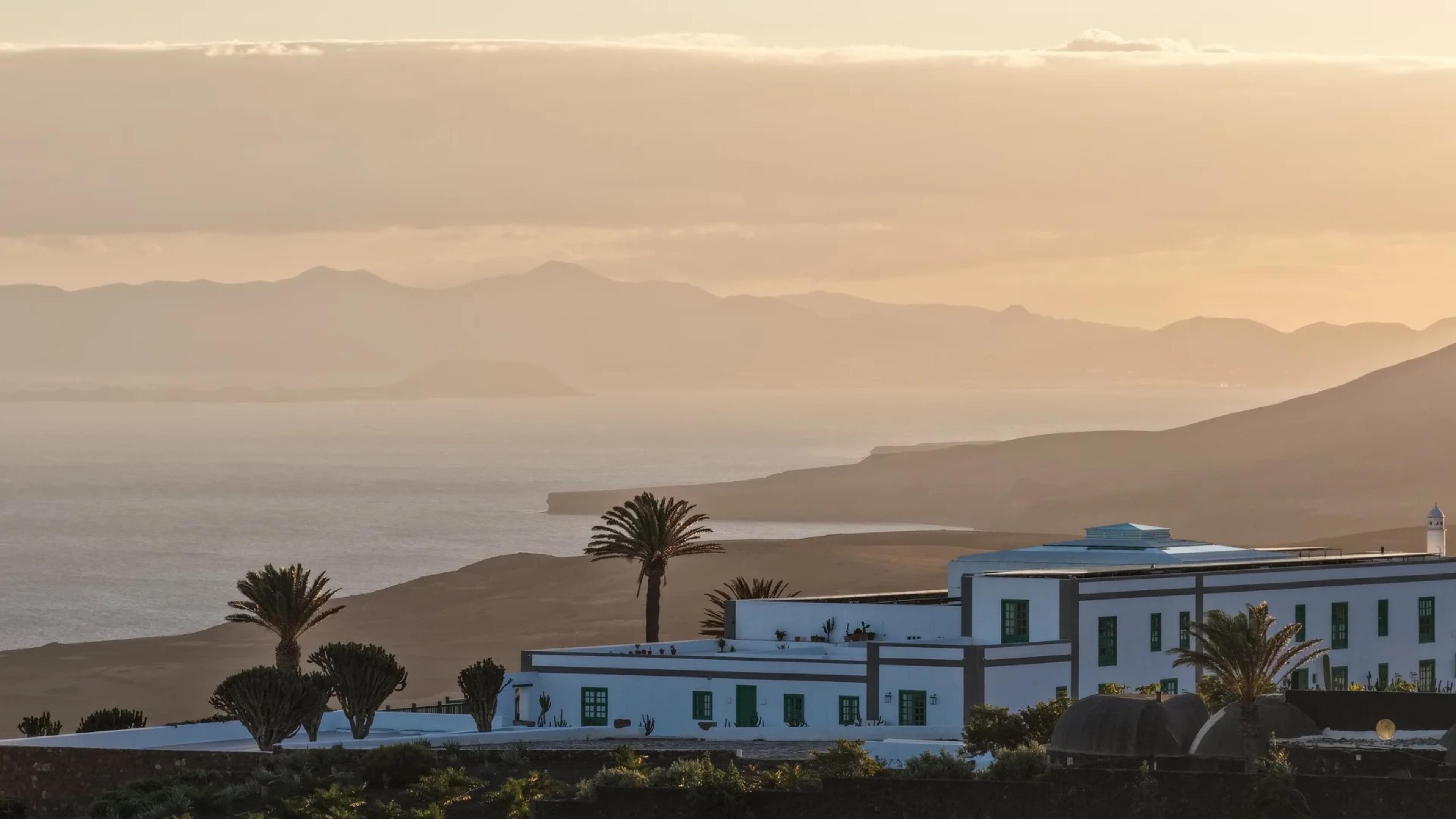 A white hotel building on a headland above the sea at sunset, with palm trees in front and the coastline and distant hills fading into haze behind it.