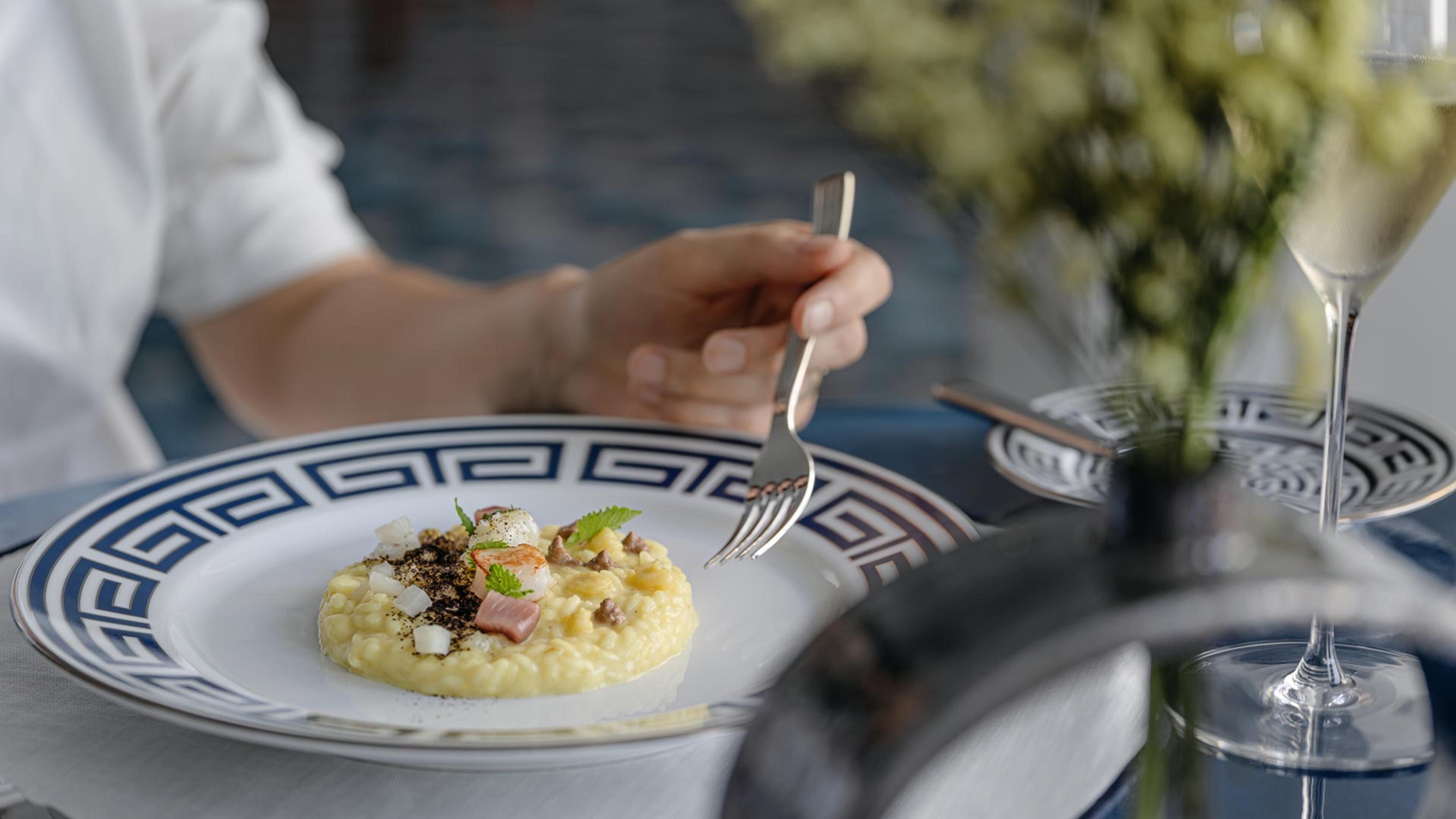 A plated risotto with ham and herbs on a blue and white patterned dish, with a diner's hand holding a fork, a flower arrangement and a wine glass out of focus in the foreground.