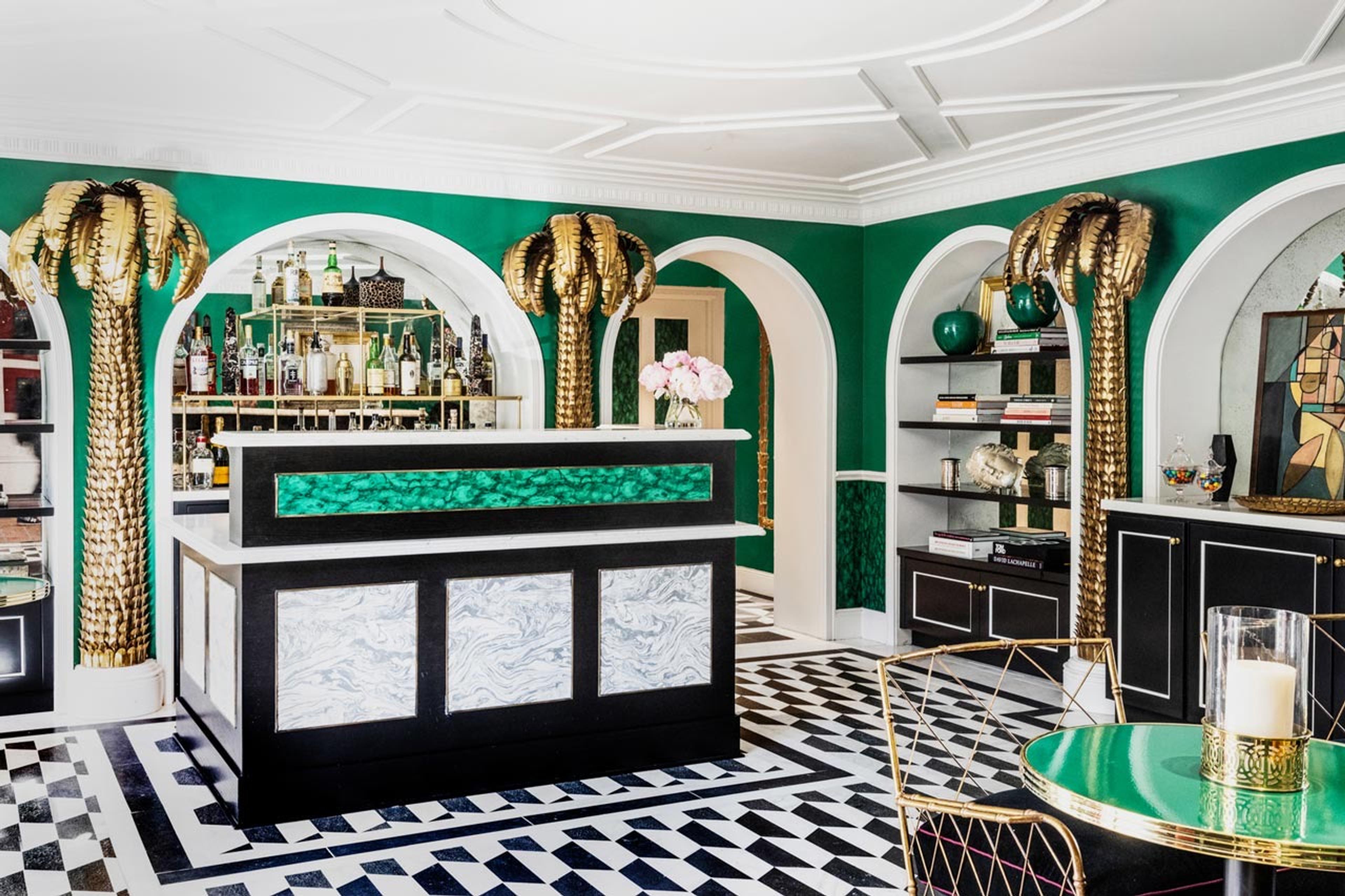 A green walled bar and lounge with gold palm tree sculptures either side of the bar, bookshelves, and a black and white checkerboard marble floor.
